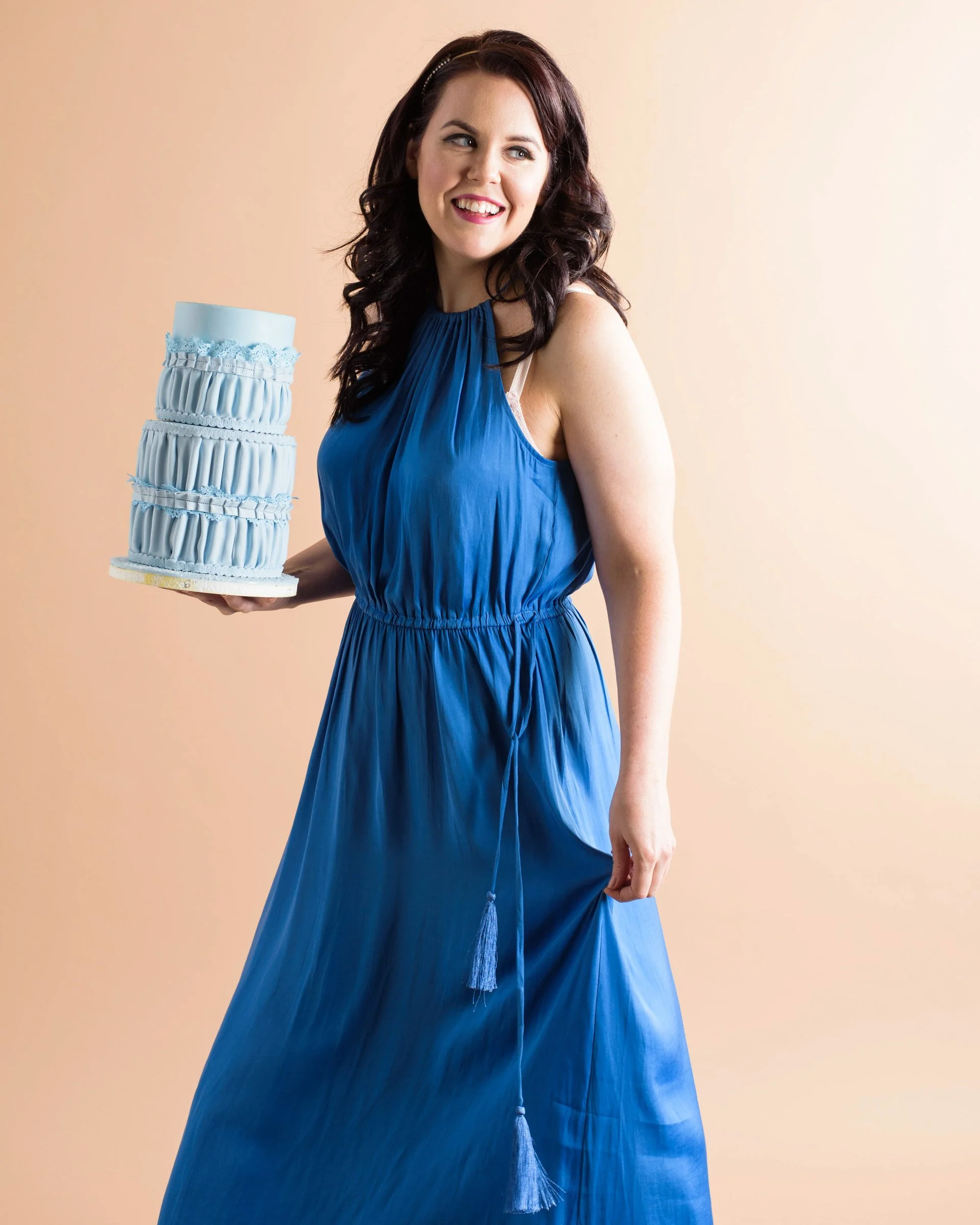 A woman with dark brown, wavy hair in a blue dress holding a tiered blue cake with ruffled and lace details, smiling and looking to the side, against a beige background.