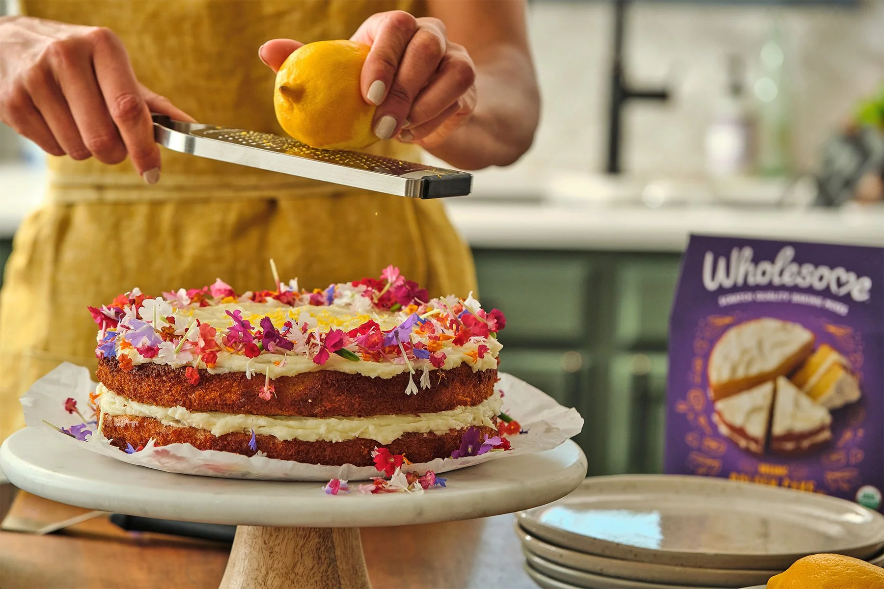 Person grating lemon zest over a layered cake decorated with edible flower petals on a cake stand.