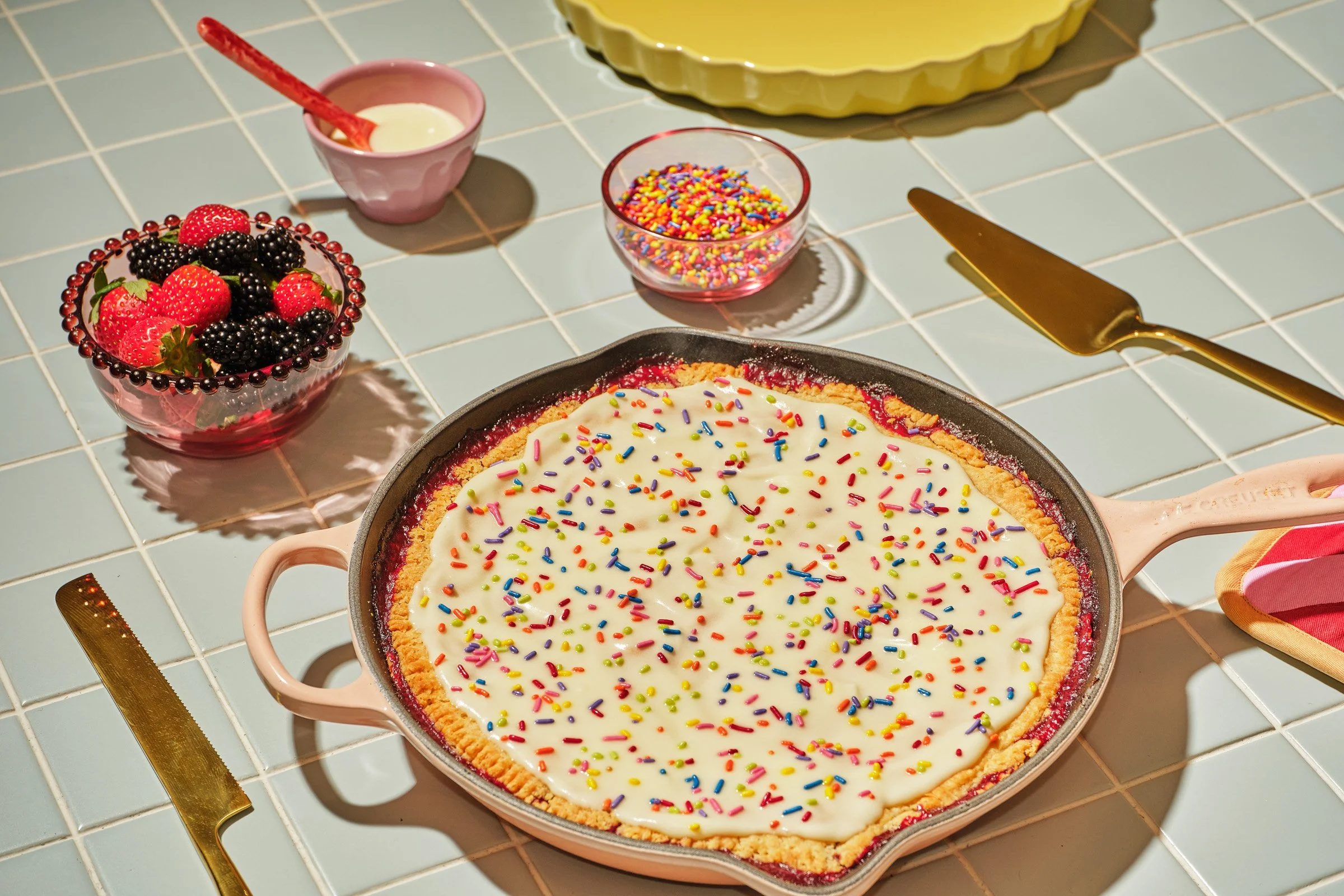 A fruit tart with colorful sprinkles in a baking dish on a white tiled countertop, surrounded by small bowls of various sprinkles, a knife, and a gold cake server.