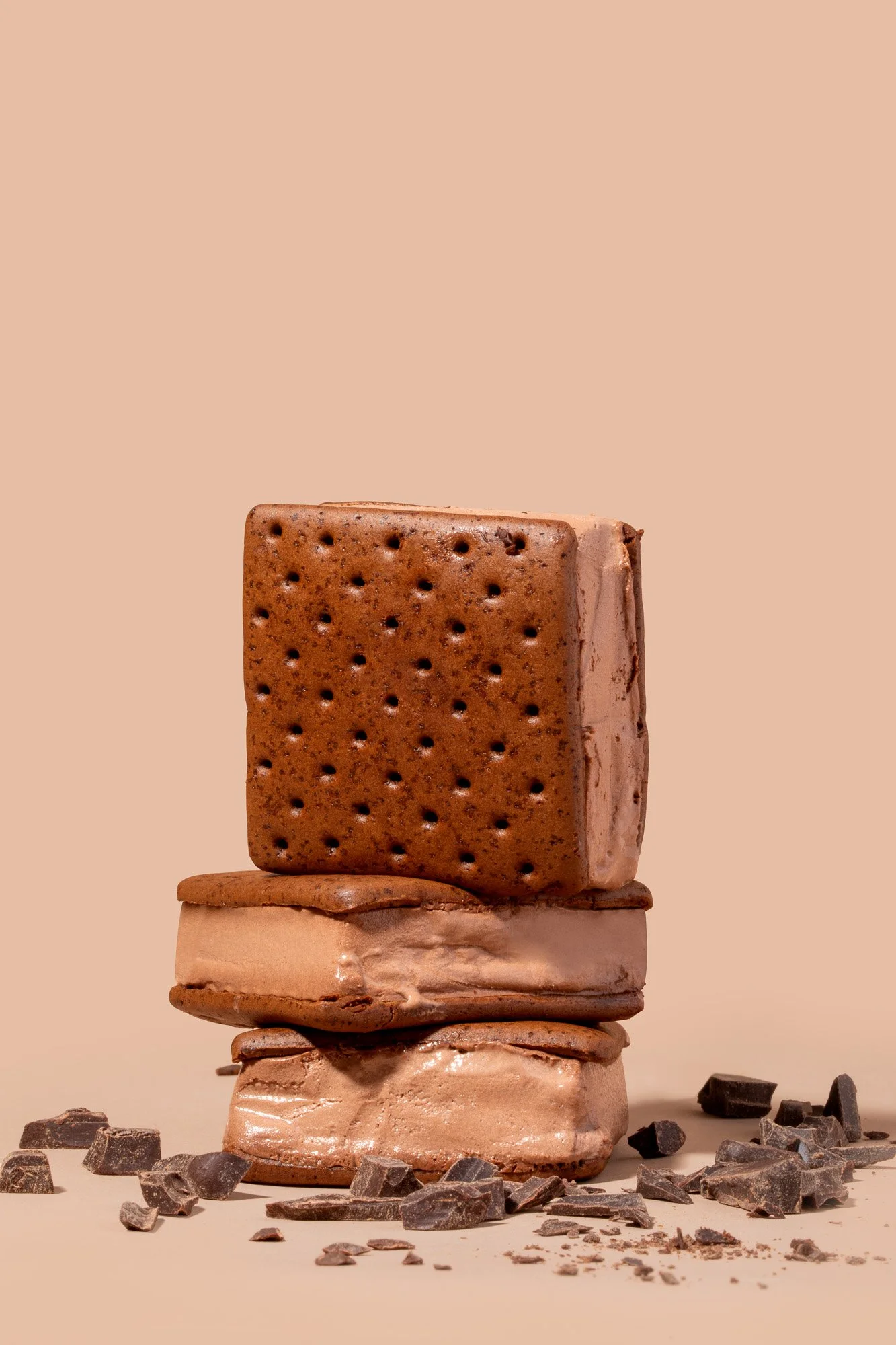 A stack of chocolate ice cream sandwiches with broken chocolate pieces scattered around on a beige background.