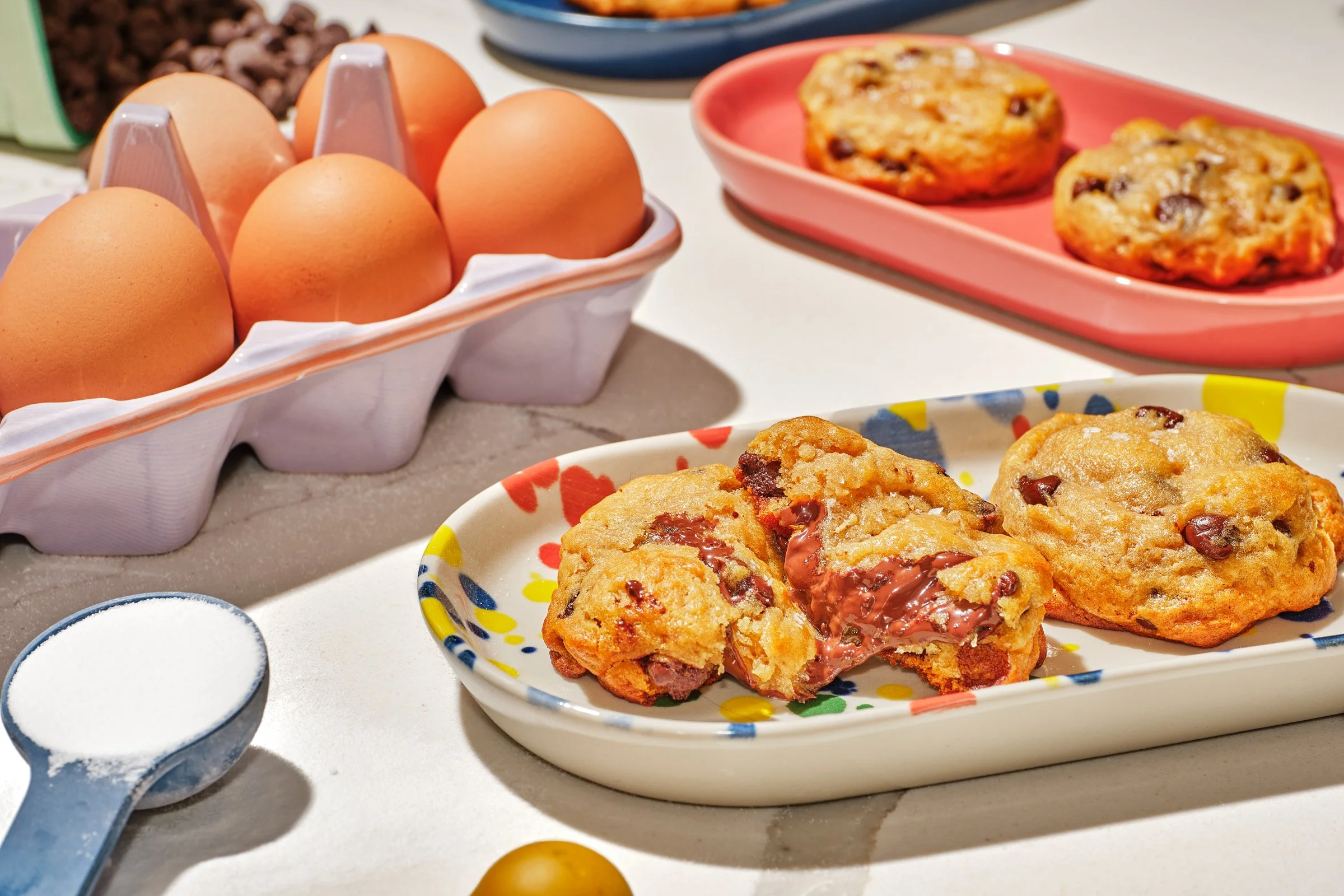 A plate of chocolate chip cookies, a tray of brown eggs, a bowl with sugar, and various baking ingredients on a kitchen countertop.