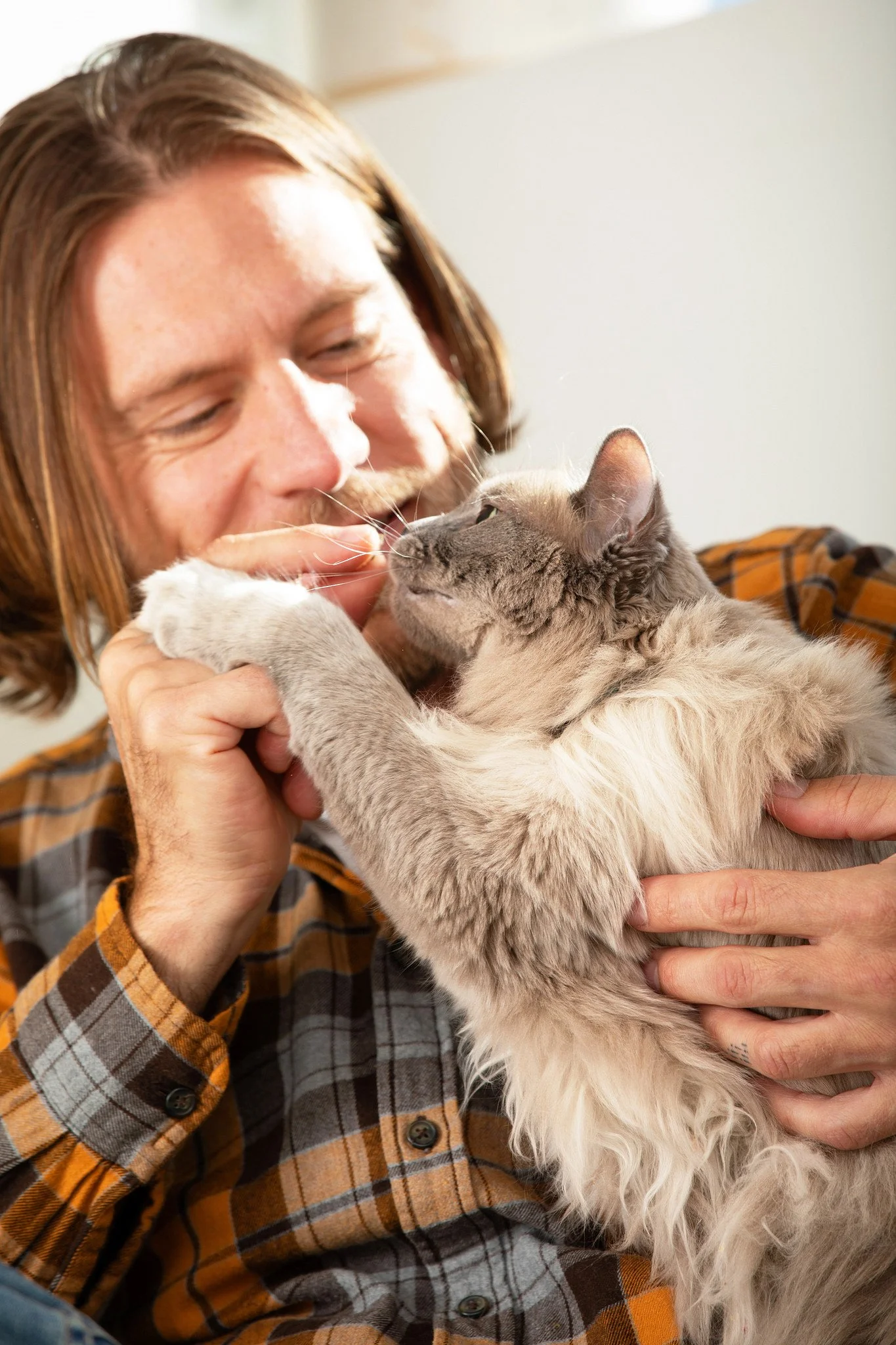 A man with long reddish-brown hair, smiling, holding and cuddling a fluffy light gray cat with darker markings, both appearing happy and affectionate.