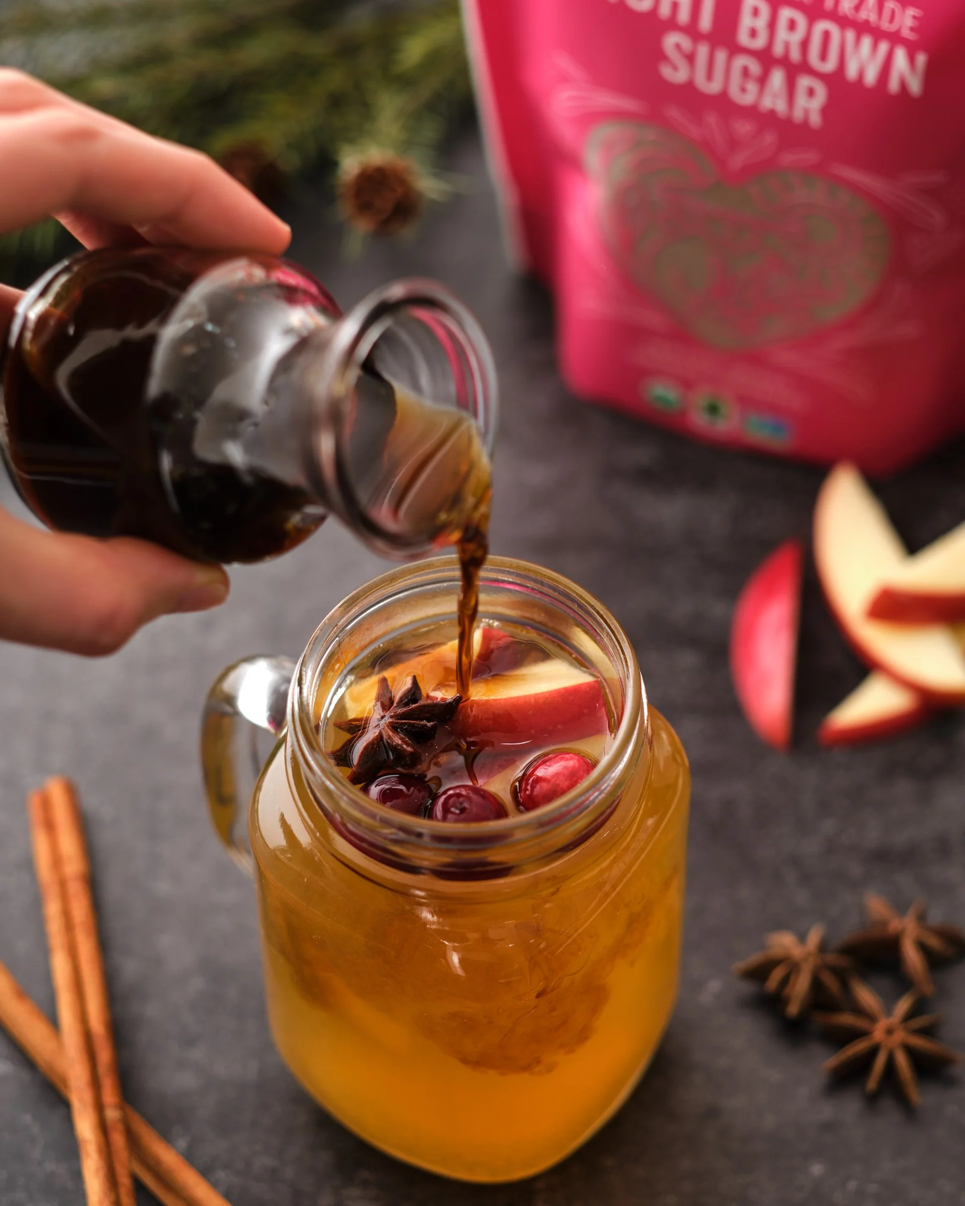 A hand holding a small jar pouring brown liquid into a glass with apple slices, star anise, and red berries on top, with a pink sugar bag and star anise on the dark surface.