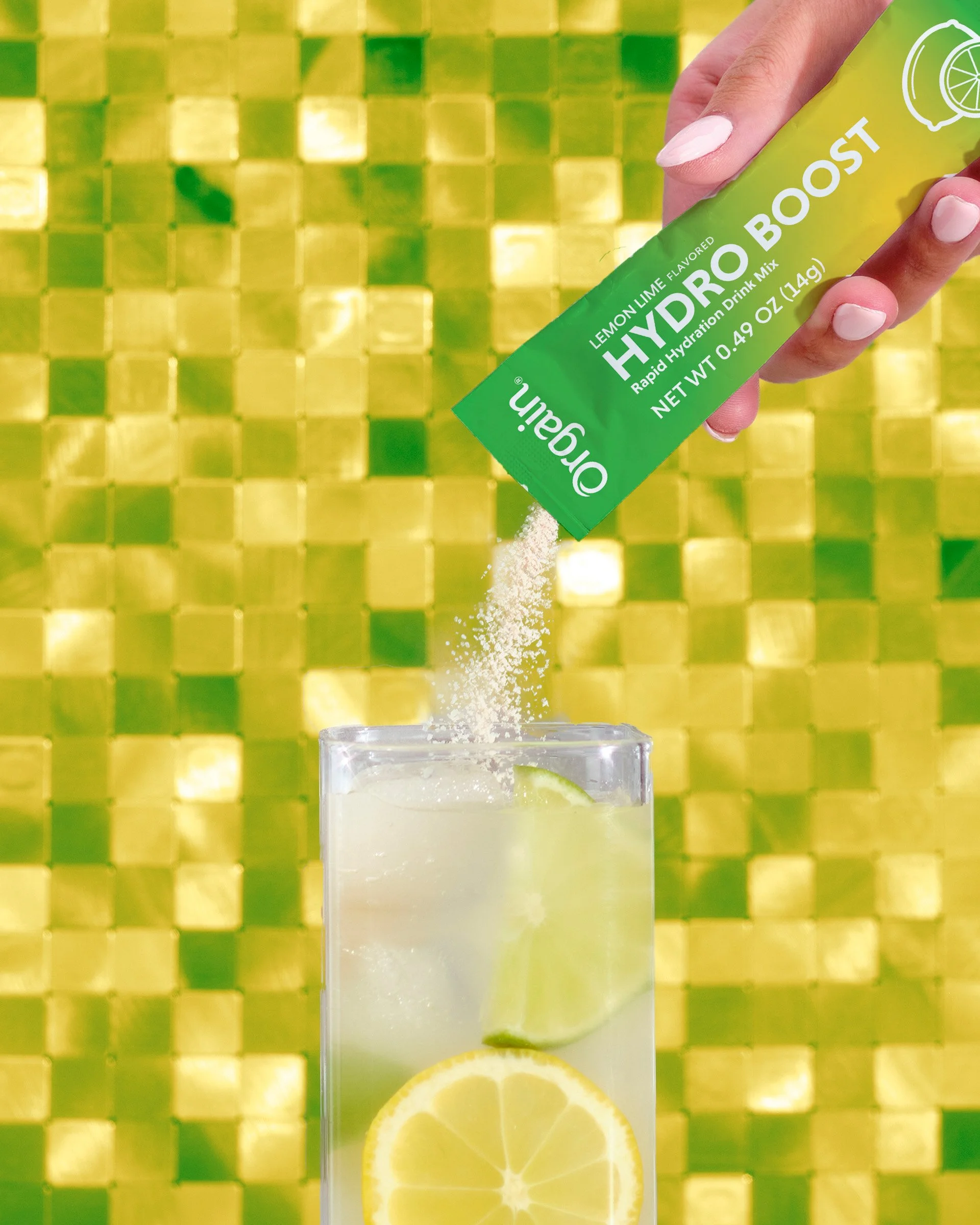 A hand holding a green packet labeled "Hydro Boost" pouring powdered lemonade flavored hydration drink mix into a clear glass with lemon slices inside, set against a shiny, yellow tiled background.