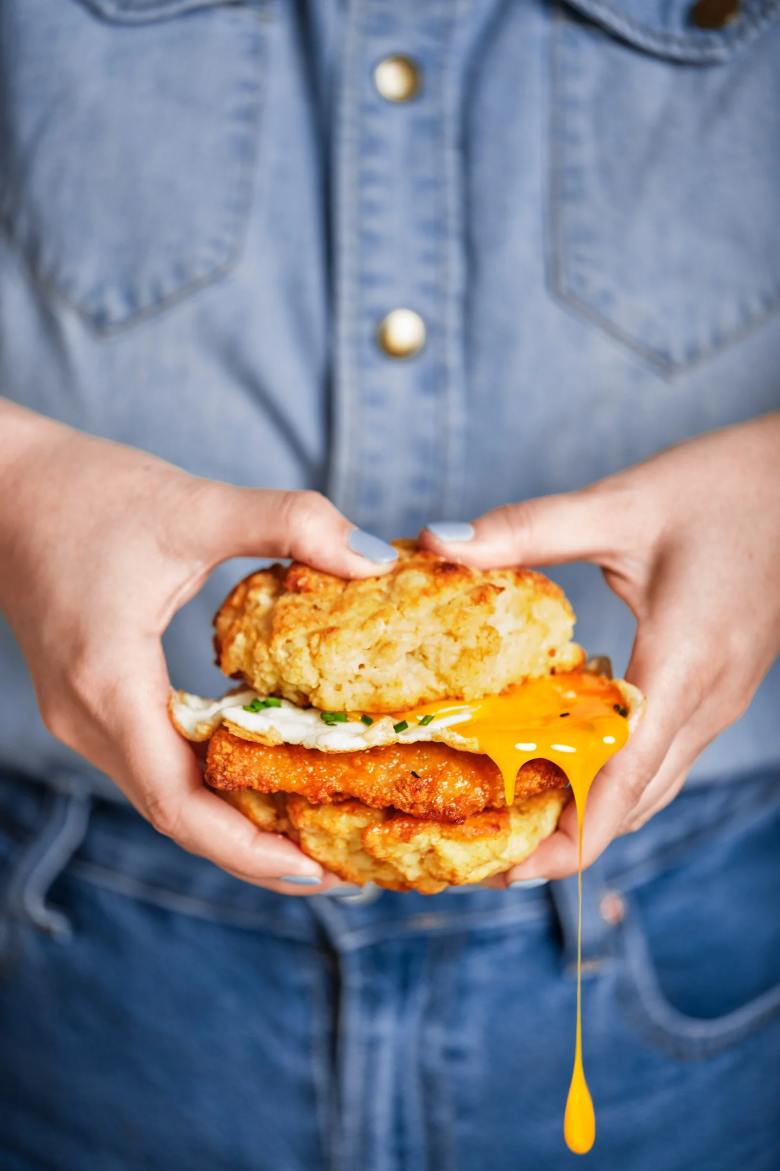 Person in a denim shirt holding a large stack of fried chicken, fried egg, and a hash brown, with yellow egg yolk dripping.