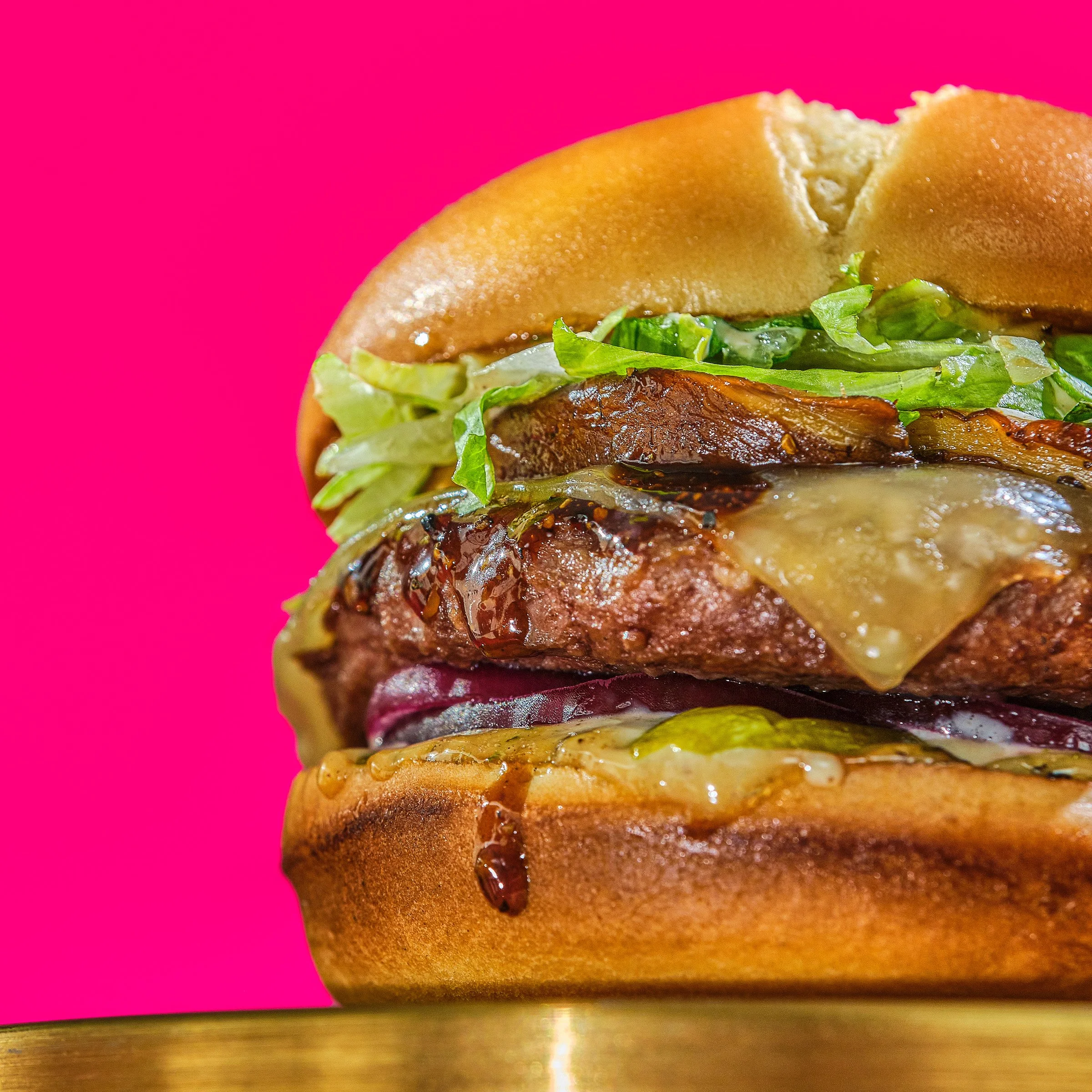 Close-up of a layered burger with lettuce, tomato, bacon, cheese, and sauce against a pink background.