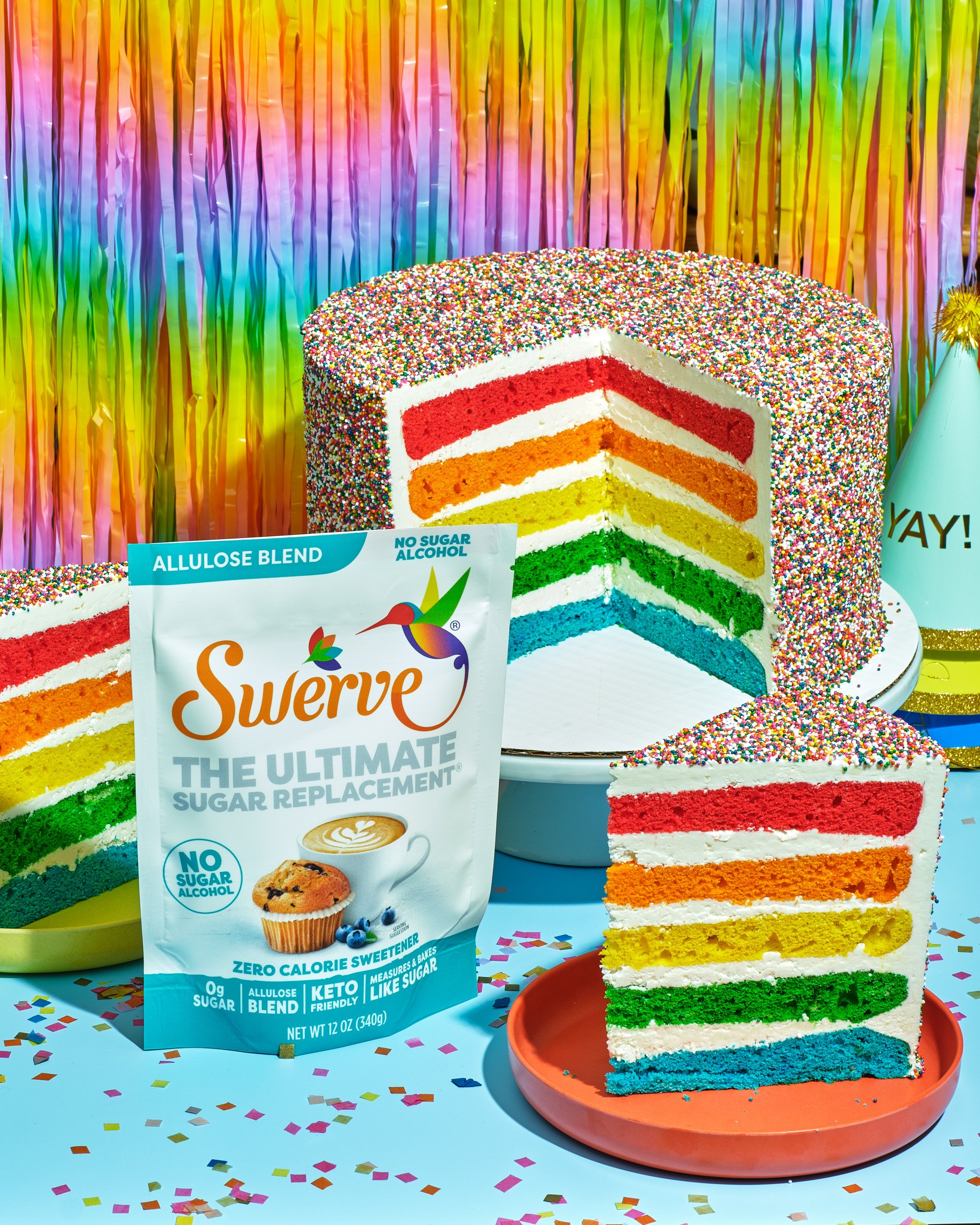 Colorful rainbow layered cake topped with multicolored sprinkles, with a slice cut out and placed on an orange plate. The background features a rainbow-colored streamers curtain, and confetti is scattered on the table around.