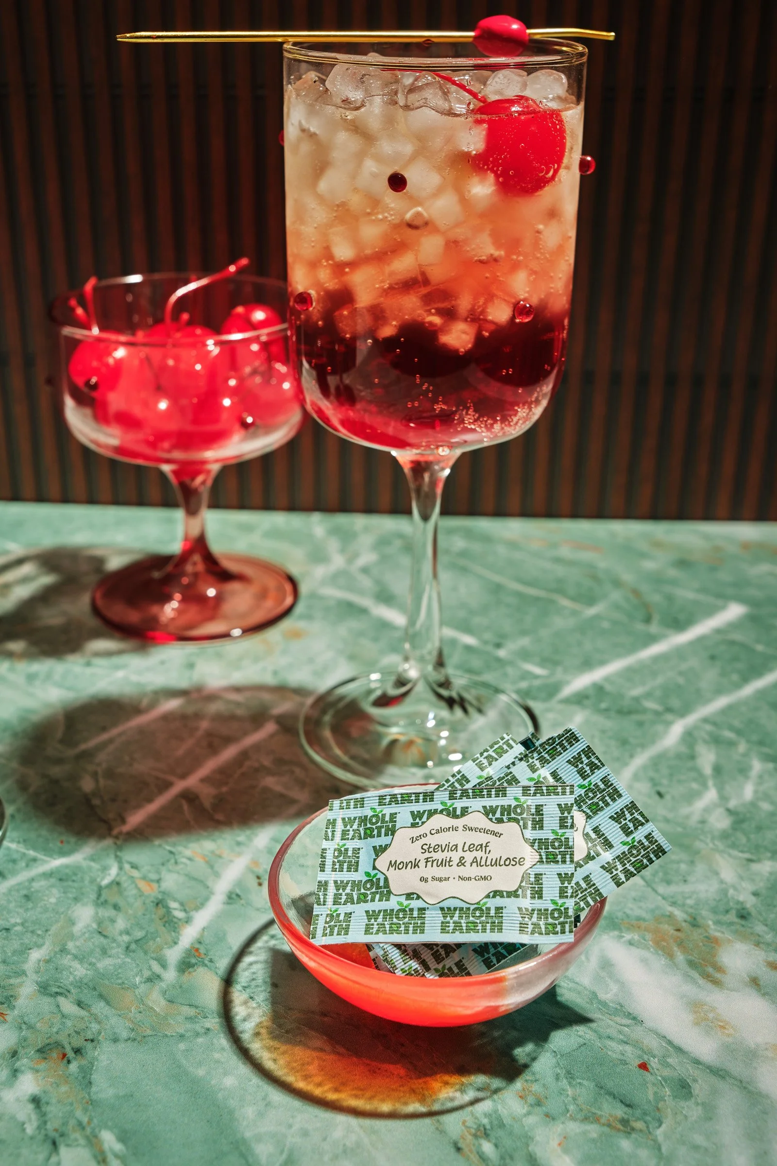 A clear glass filled with ice, maraschino cherries, and a red dessert layered on the bottom, topped with a cherry and a skewer. A smaller glass with cherries is beside it. There are small packets of Stevia leaf, monk fruit, and allulose sweetener lab