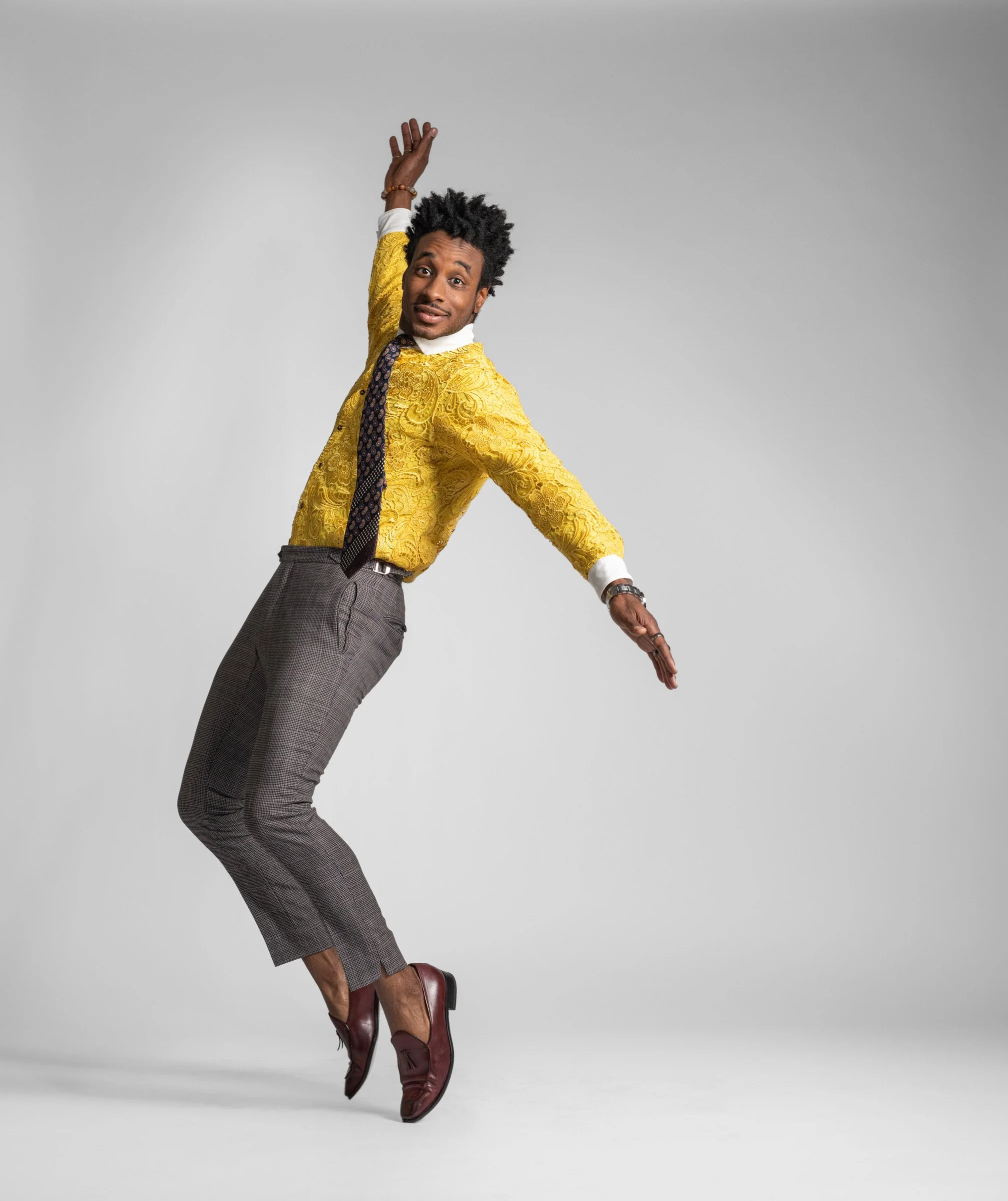 A man with a dark skin tone and curly hair is dancing or jumping against a plain gray background. He is wearing a yellow lace shirt with white cuffs and collar, a patterned tie, gray checkered trousers, and maroon leather shoes. His expression is che