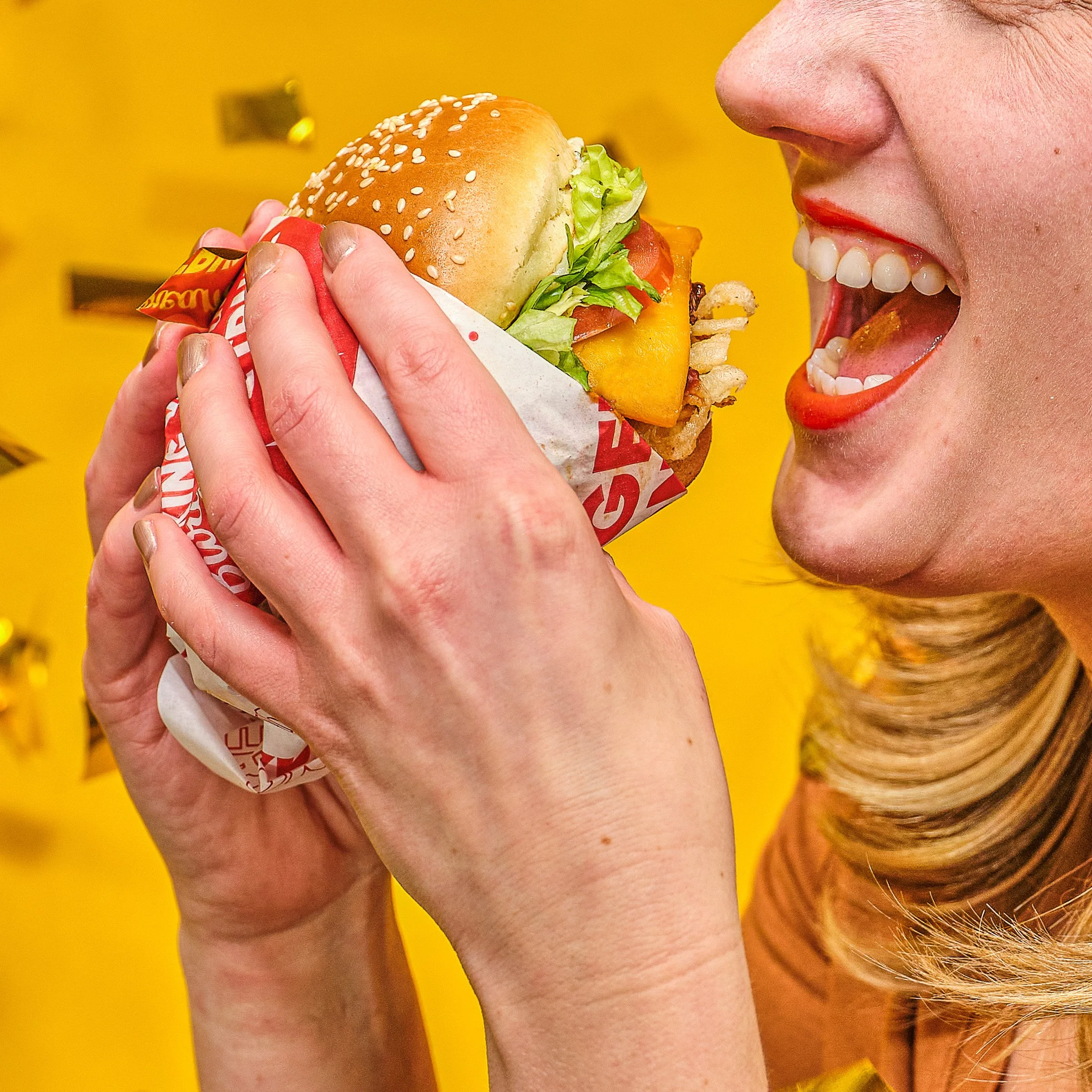 Person holding a cheeseburger with lettuce, tomato, cheese, bacon, and a sesame seed bun, smiling with an open mouth in front of a bright yellow background.