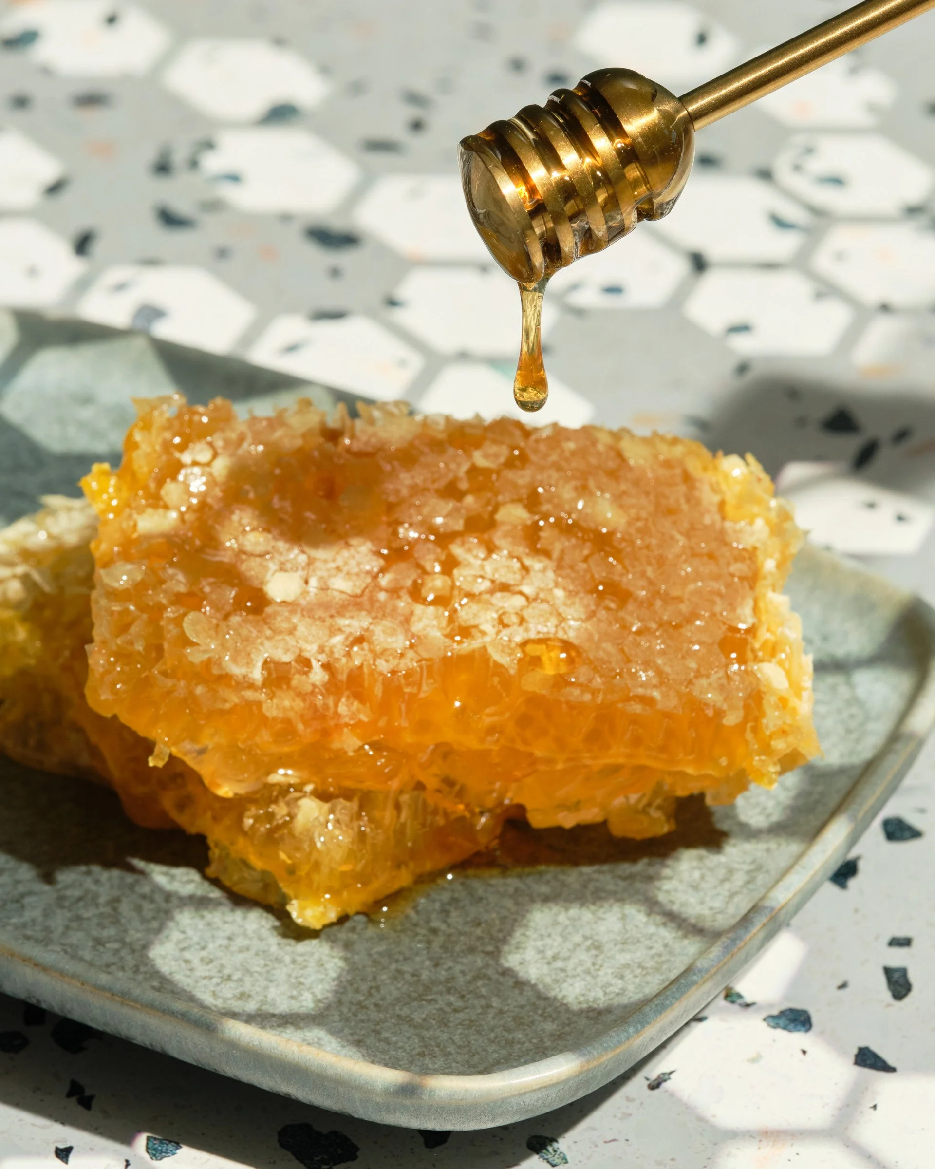 Close-up of a honey dipper dripping honey onto a piece of cake on a metal tray with a honeycomb-patterned background.