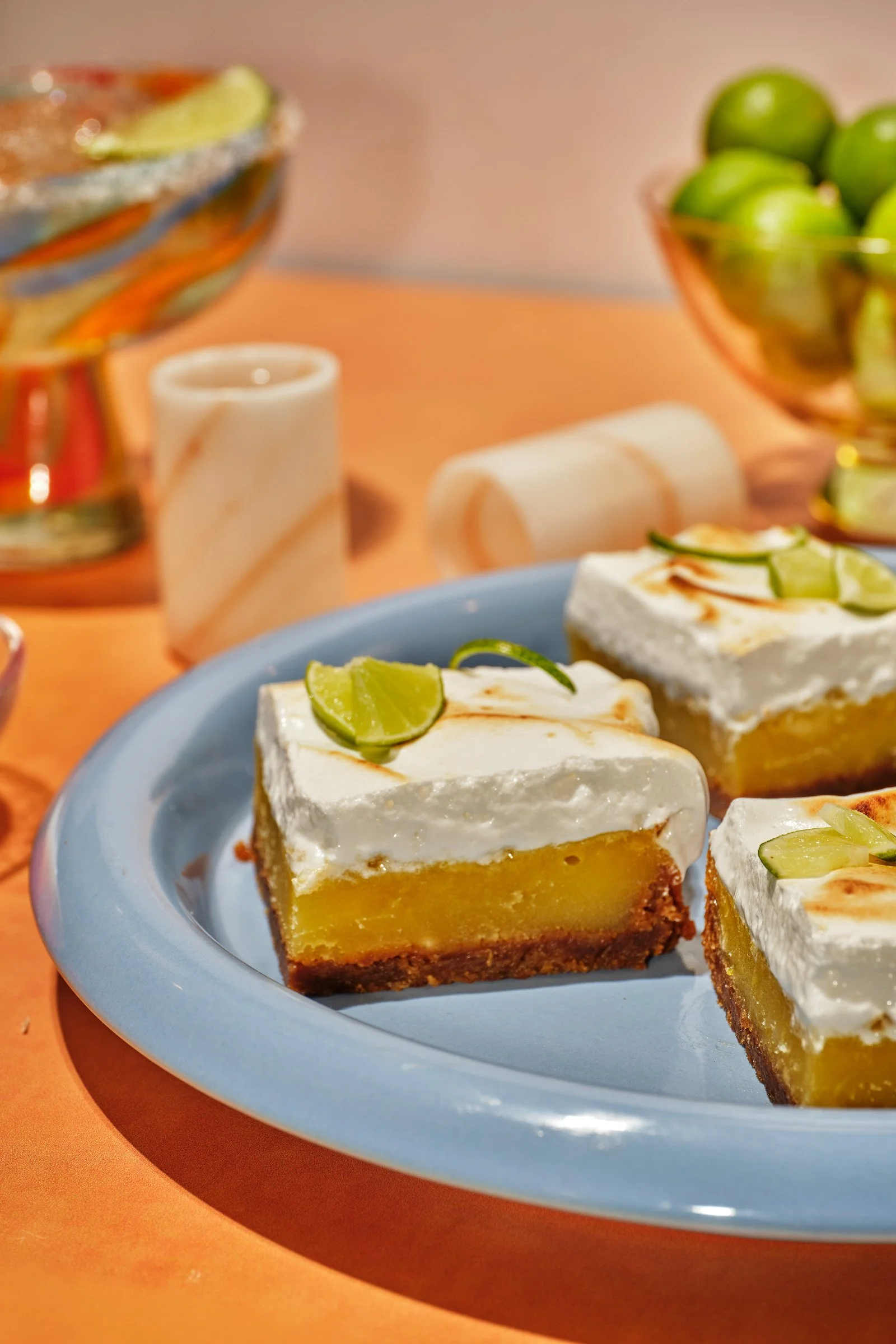 Close-up of a pieces of lime-topped lemon bars on a blue plate, with bowls of limes and a decorative bowl in the background.