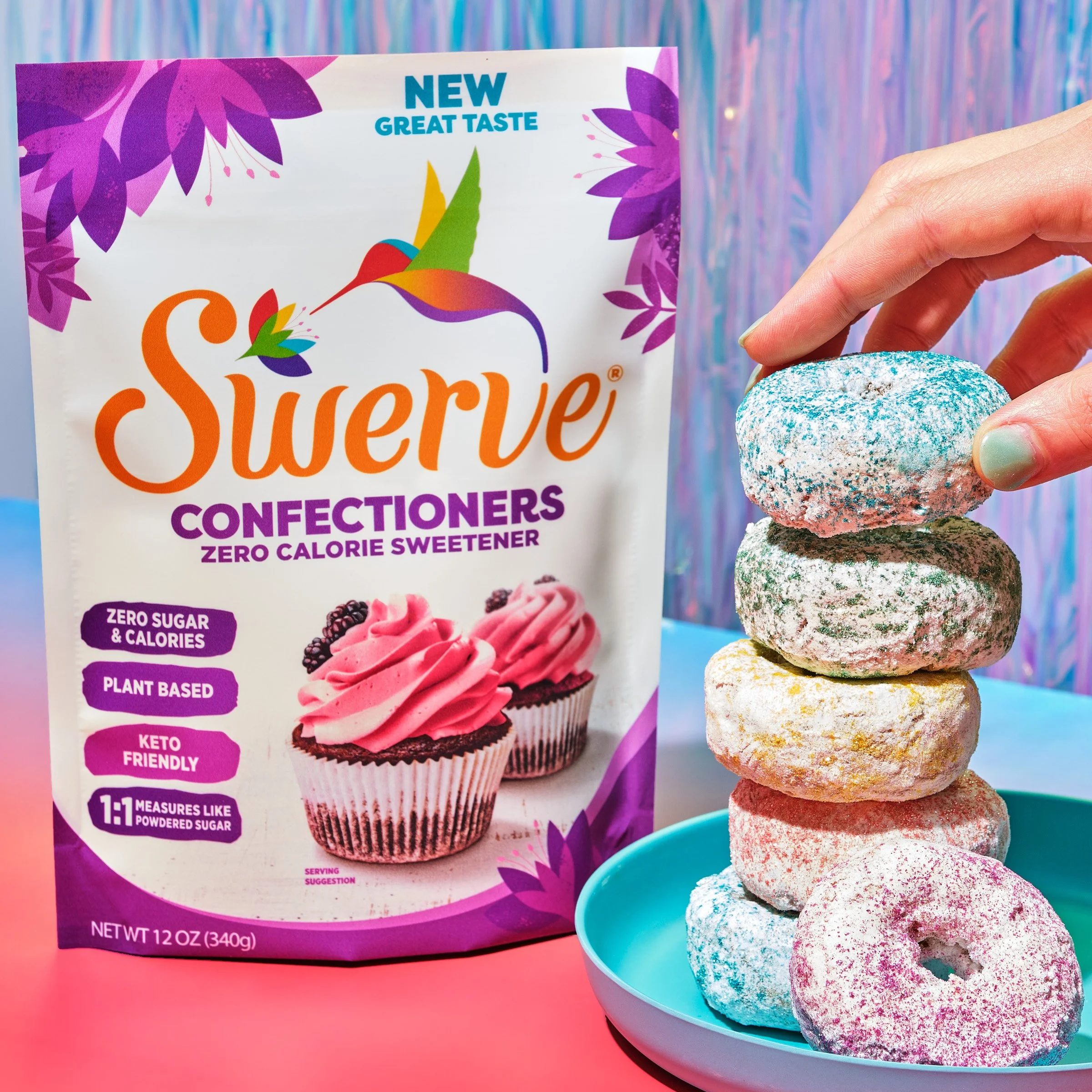 Colorful package of sugar-free confectioners with purple and pink floral design, next to a stack of colorful, speckled cookies in a light blue bowl, with a pastel-colored background.