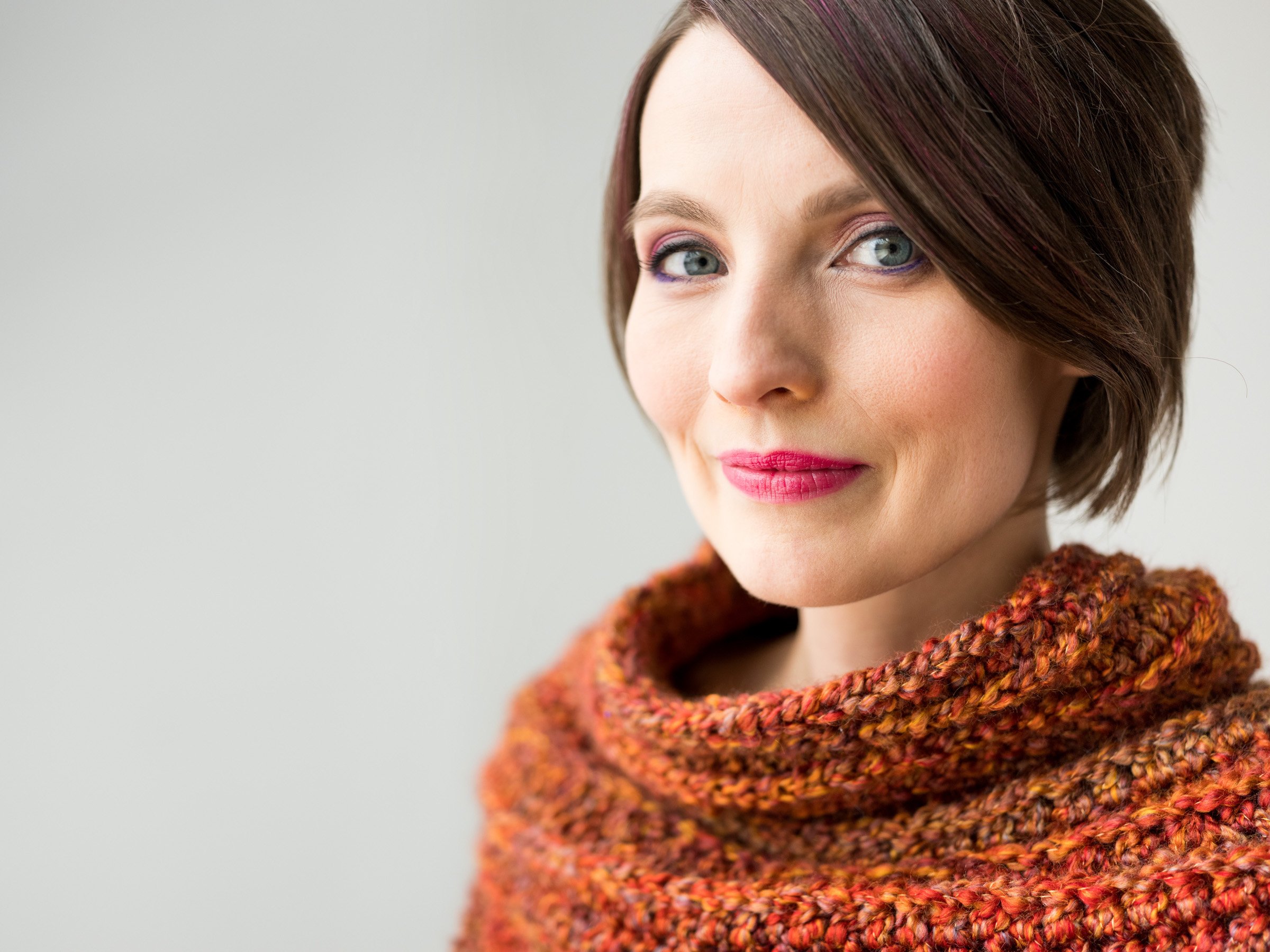 Close-up of a woman with short, tousled brown hair, wearing makeup with pink lipstick and colorful eye makeup, dressed in a multicolored knitted sweater.