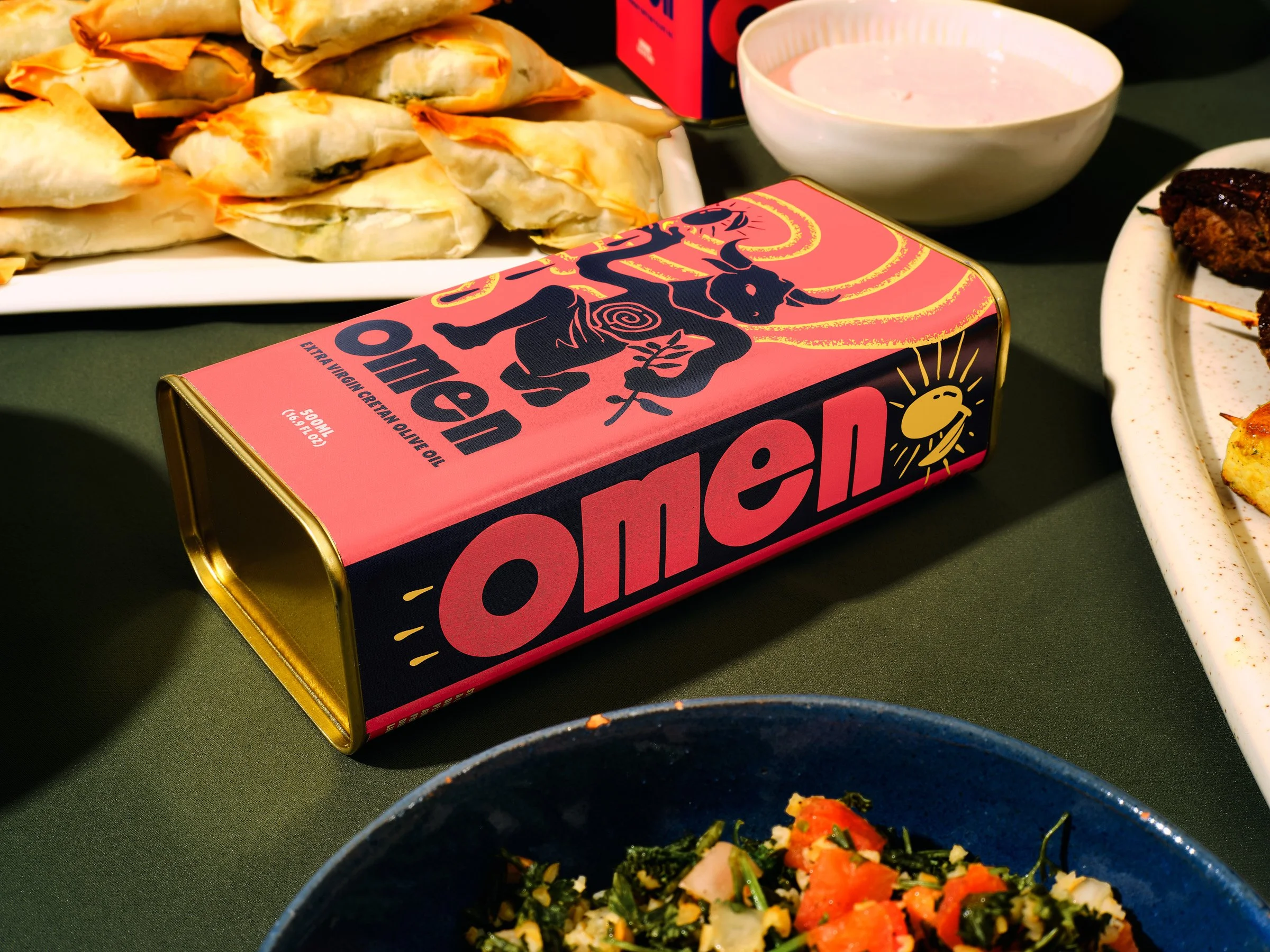 Close-up of a table with spring rolls, a bowl of pink sauce, a box of extra virgin olive oil, and a salad in a black bowl, with a plate of grilled meat on the side.
