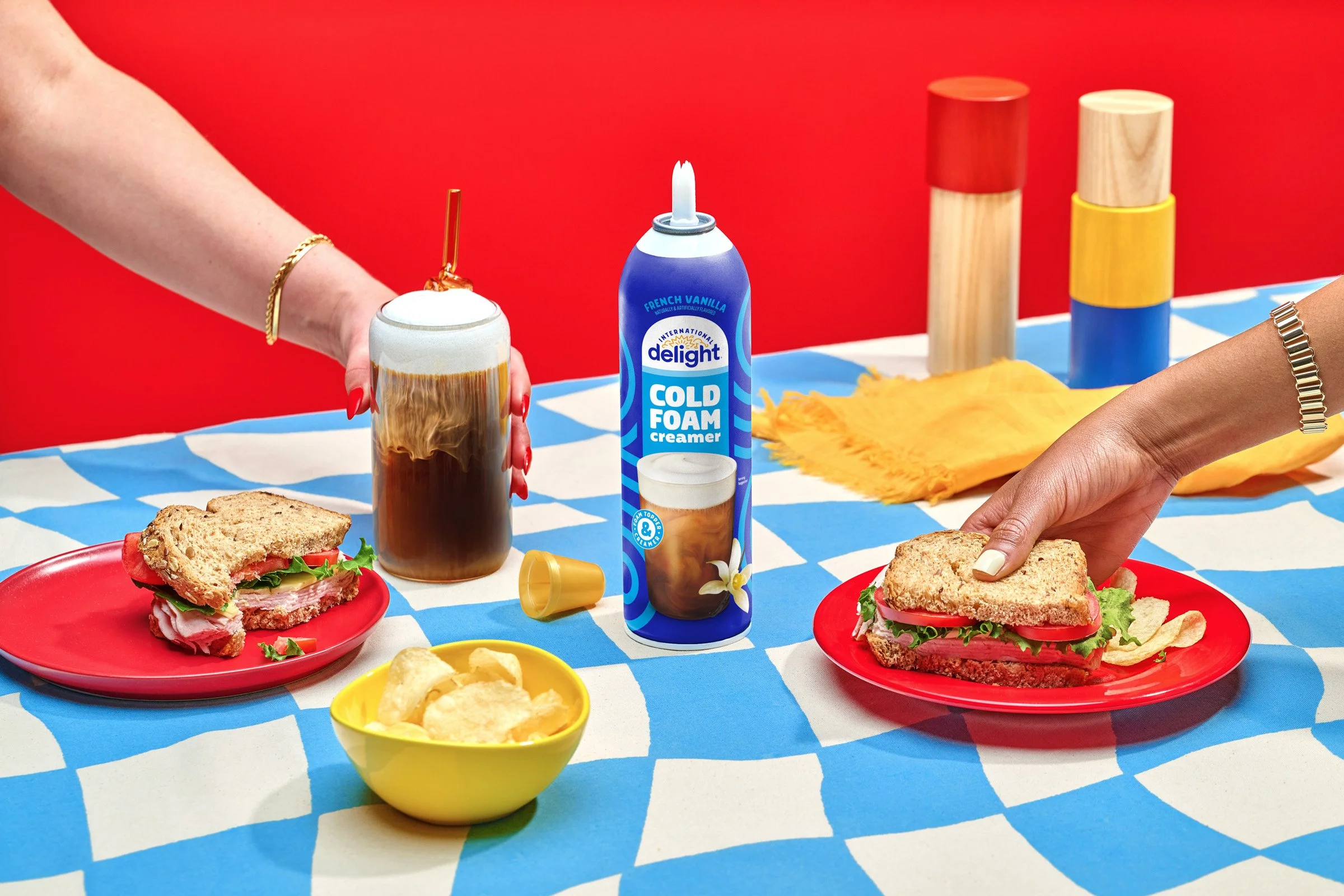 A colorful table setting with two sandwiches on red plates, a bowl of potato chips, and a can of joy foam cooling cream. There are also a slice of cake, a burger, and some chips on a yellow dish, with a backdrop of a red wall and various cylindrical 