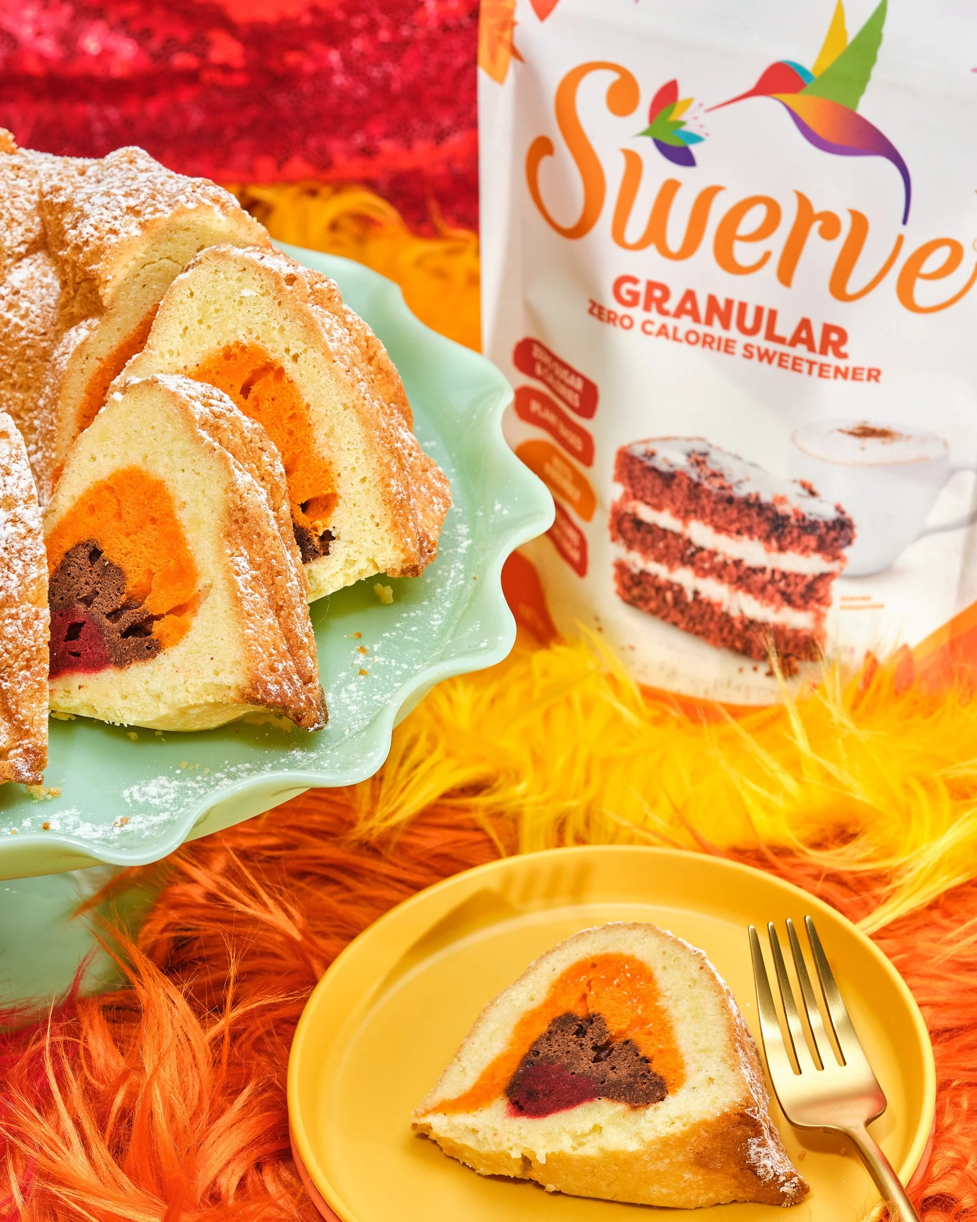 Sliced marble cake with orange and chocolate layers on a yellow plate, fork beside it; powdered sugar sprinkled on top; package of sweetener powder visible in the background, colorful surface with orange feathers.
