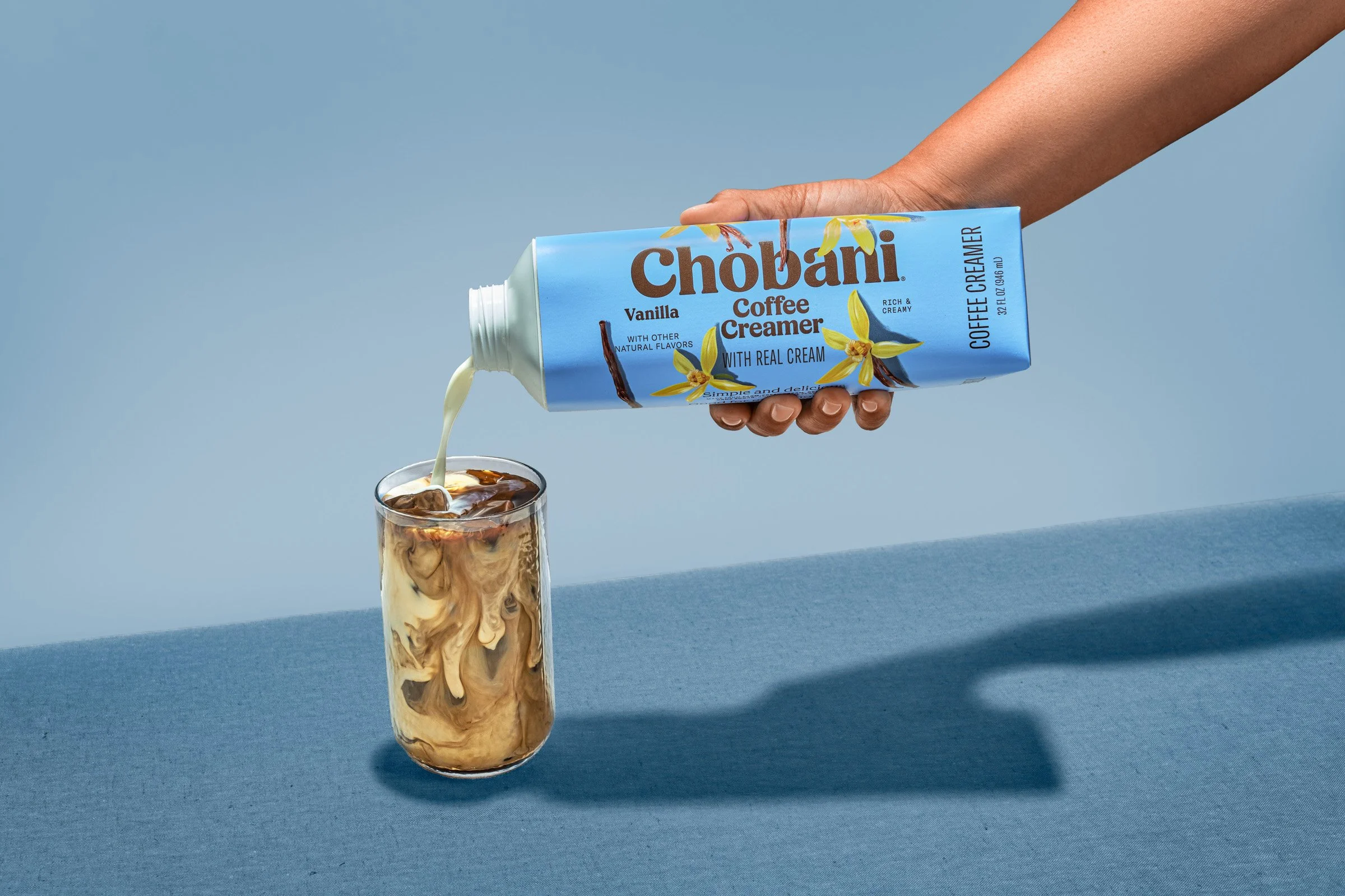 A hand holding a blue carton of Chobani vanilla coffee creamer over a glass of iced coffee with swirled cream.