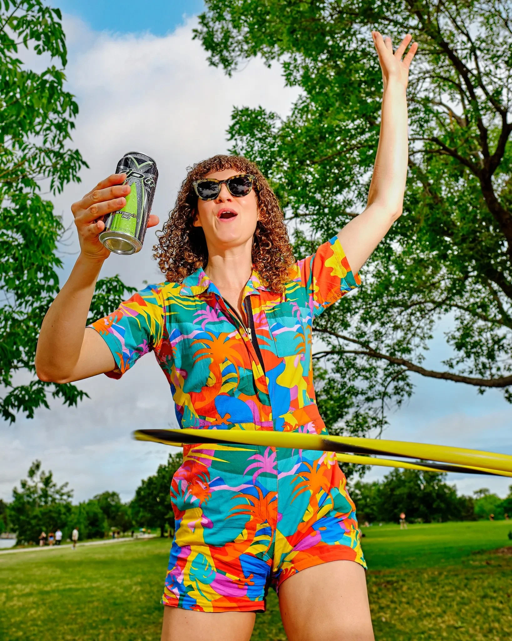 A woman with curly hair wearing colorful tropical print outfit and sunglasses, holding a can, playing hula hoop in a park on a sunny day.