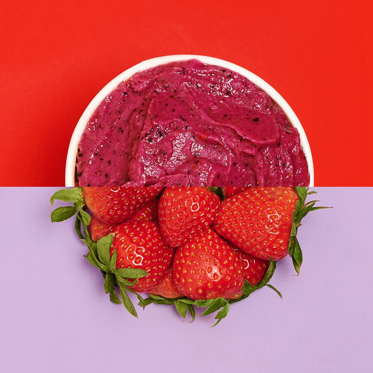 A bowl of purple berry ice cream or sorbet placed above a bunch of fresh strawberries on a bisected background of red on the top and light purple on the bottom.
