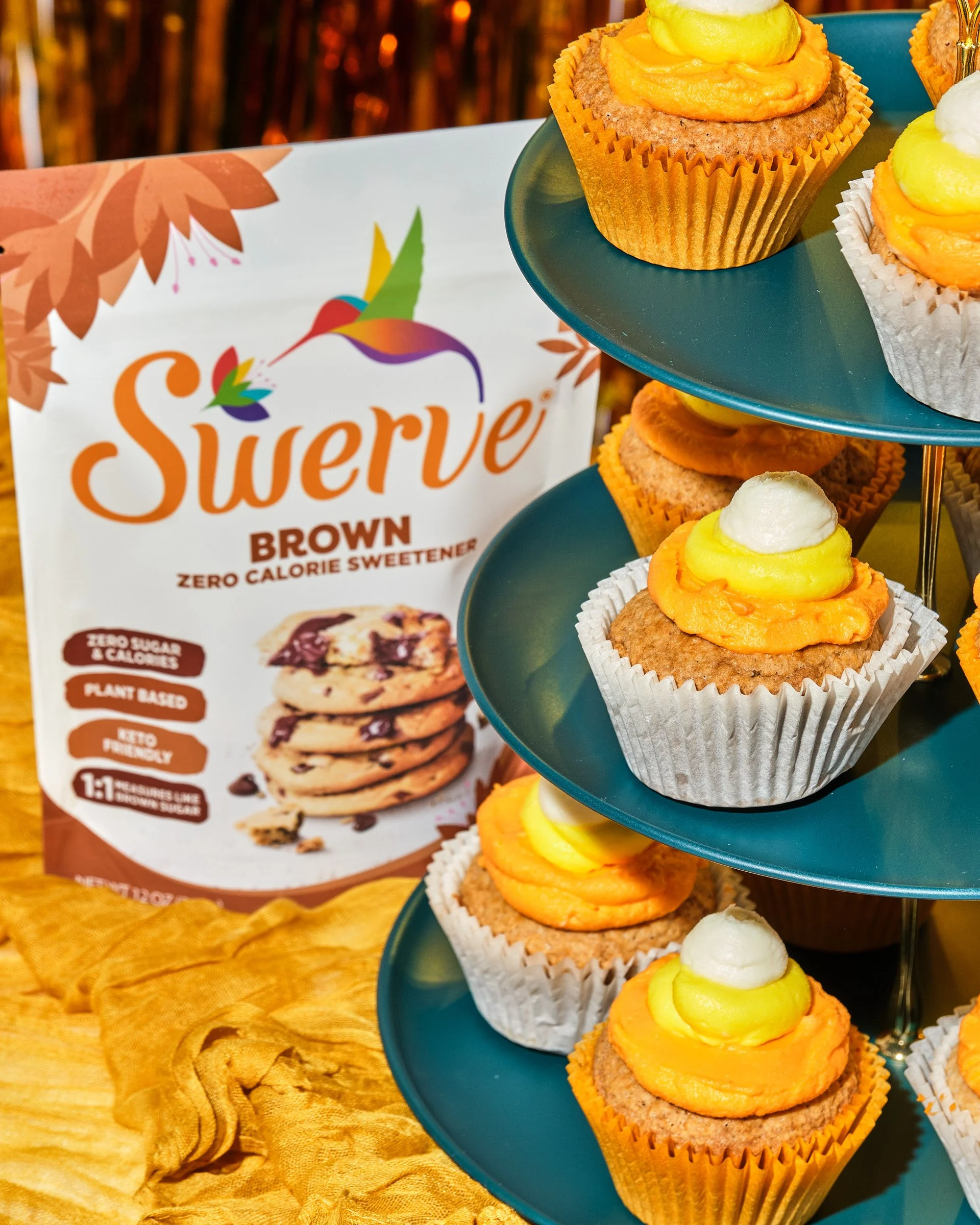 Close-up of a tiered stand with cupcakes topped with yellow and white frosting. A sign in the background displays a logo for Swerve Brown, indicating the product may be a zero-calorie sweetener.