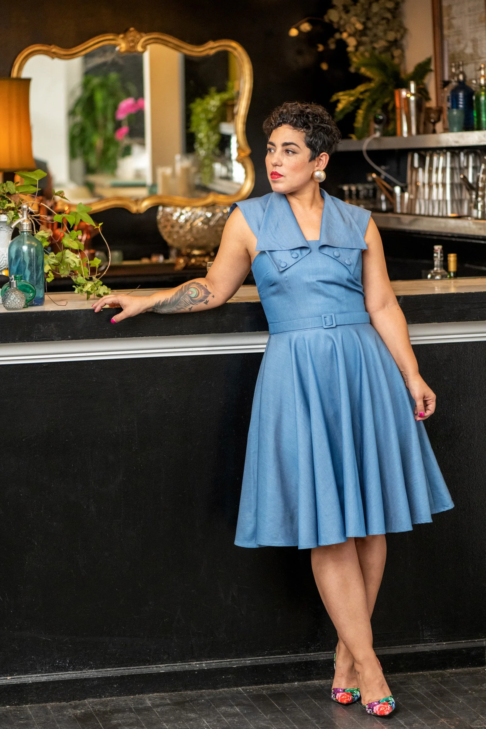 A woman with short curly hair and tattoos on her left arm leaning against a black bar counter, wearing a sleeveless blue dress with a belt and colorful floral high heels, in a stylish bar setting with plants, bottles, and an ornate mirror in the back