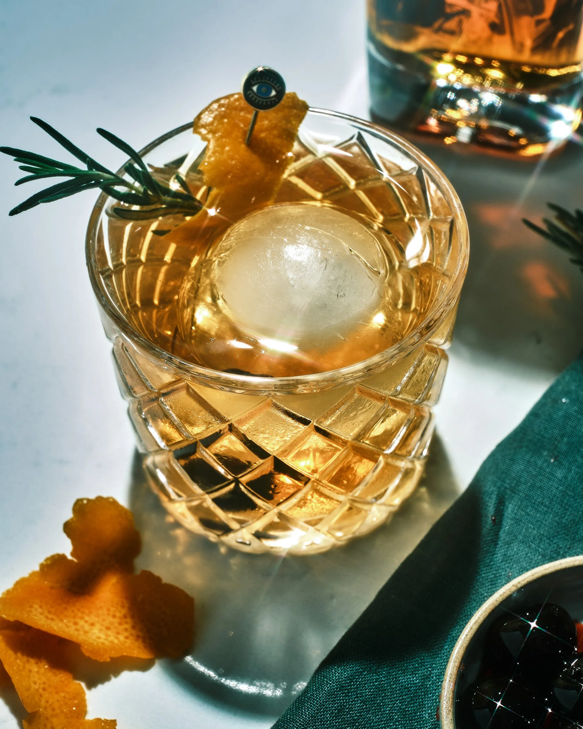Close-up of a glass with ice and a golden-colored beverage, garnished with a sprig of rosemary and a lemon peel, placed on a white surface with some cookies nearby, and another glass in the background reflecting light.