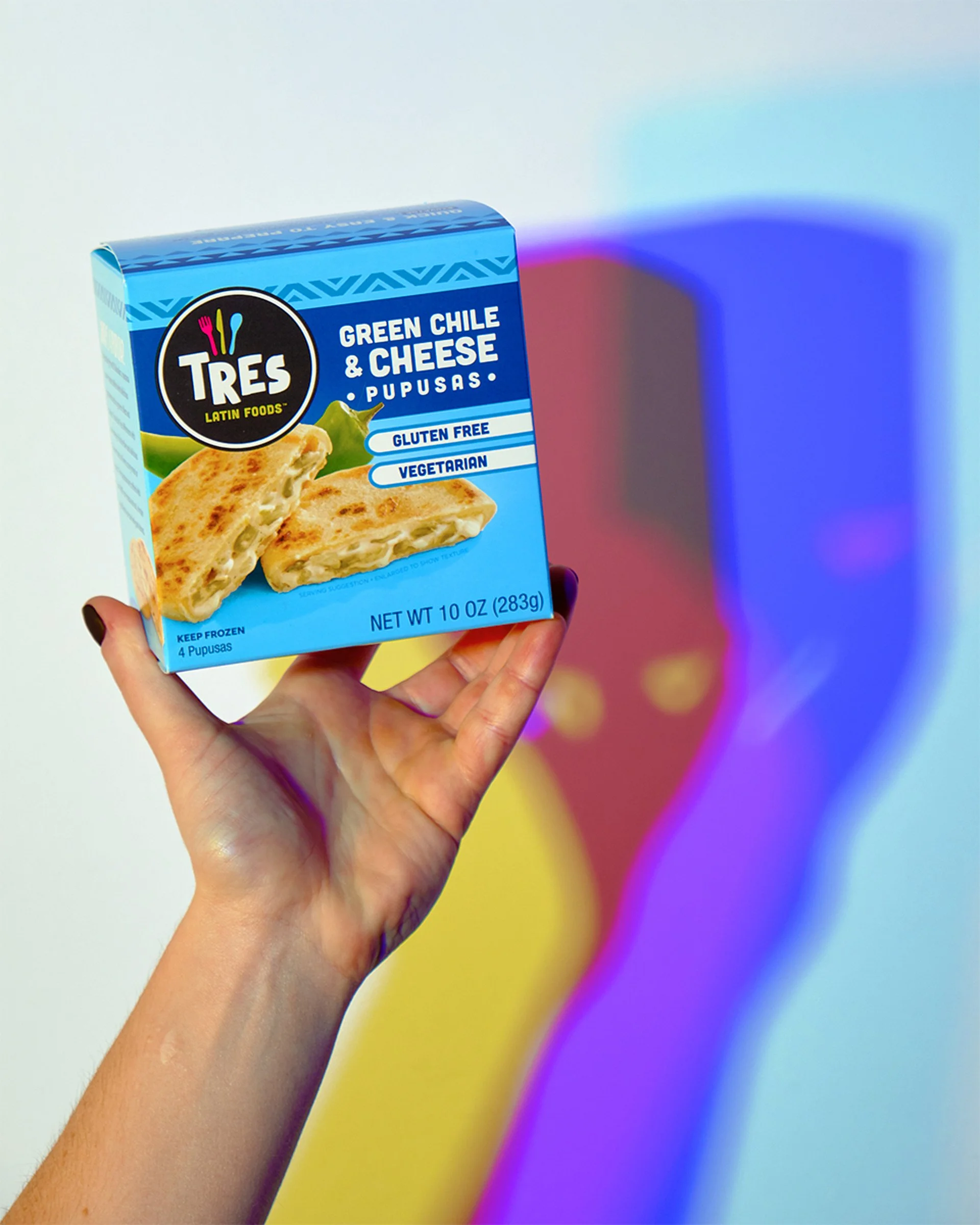 A hand holding a blue box of Tres Latin Foods Green Chile & Cheese Pupusas against a colorful blurred background.