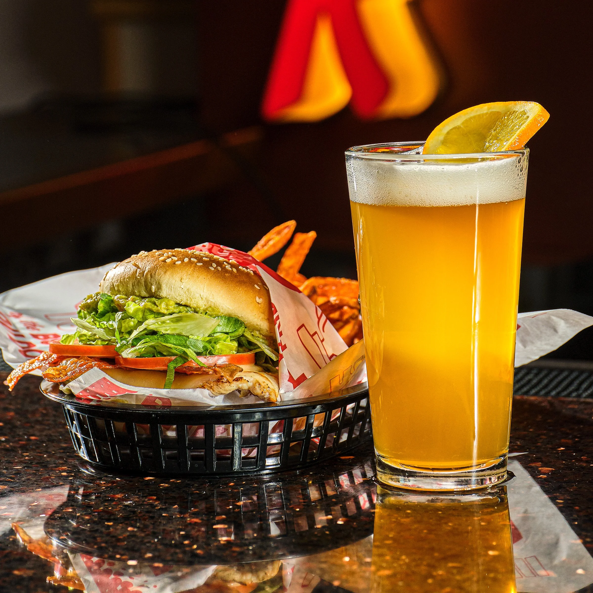 A fast food meal includes a sandwich with lettuce, tomato, and chicken in a bun with sesame seeds, some fries, and a glass of orange juice with a lemon wedge, on a dark granite countertop.