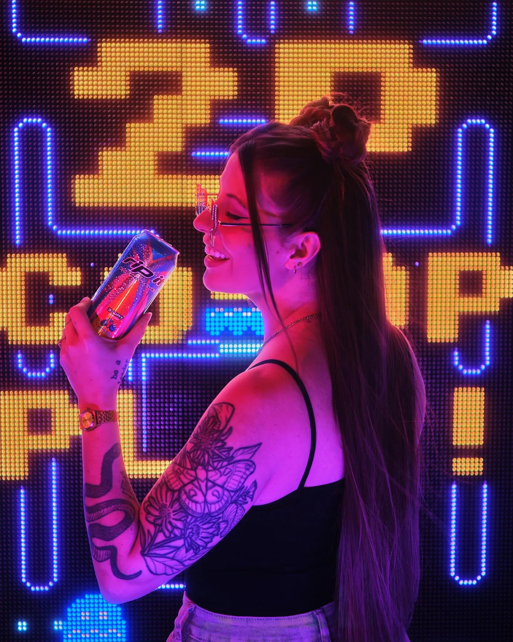 A woman with long dark hair and tattoos on her arm smiling while holding a colorful candy bar in front of an illuminated pixel art background.