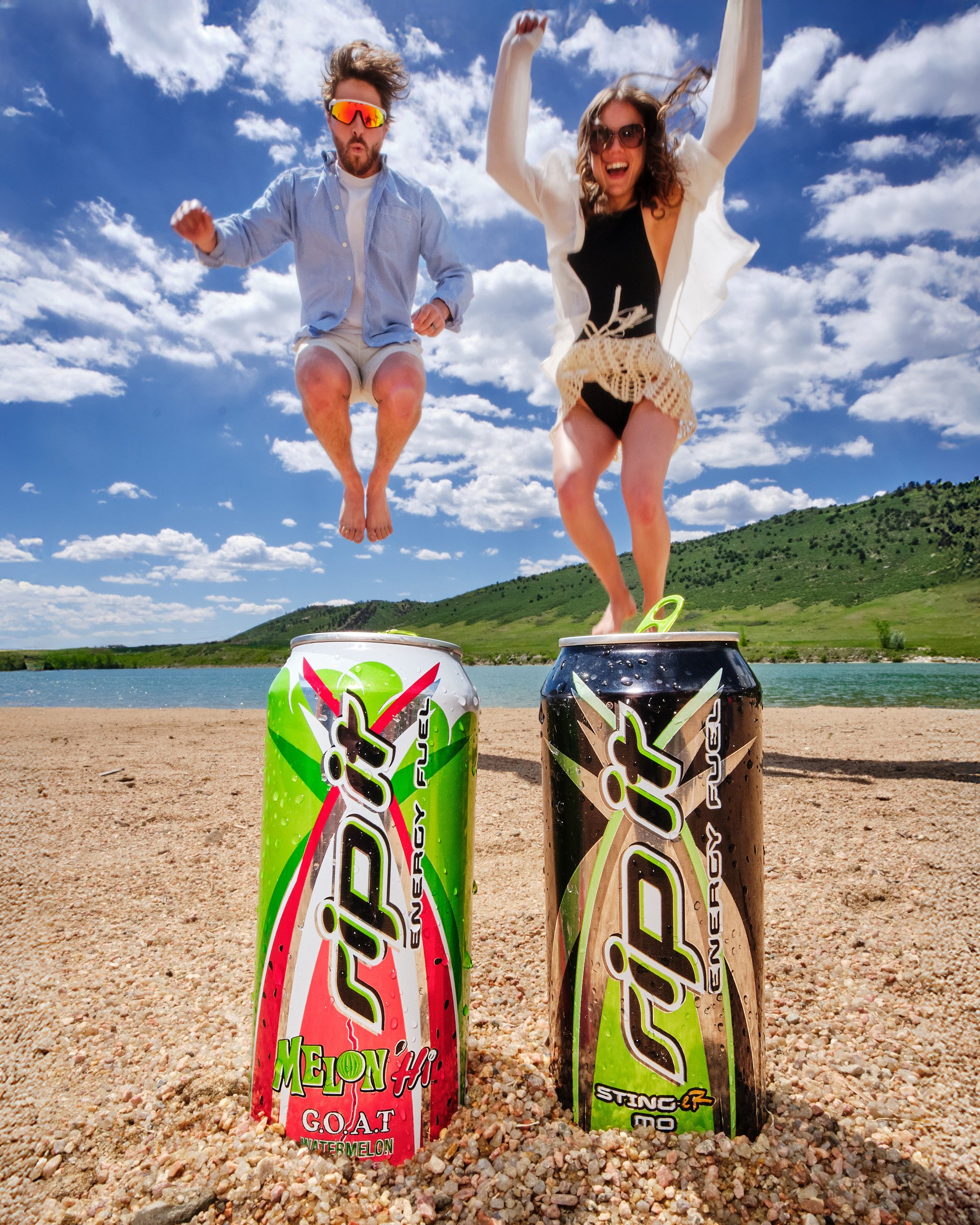 Two young people jumping on rocky ground by a river with a green, hill-covered landscape, under a blue sky with white clouds. One man with sunglasses and a beard, wearing a blue shirt and shorts, and one woman with sunglasses, wearing a white cardiga
