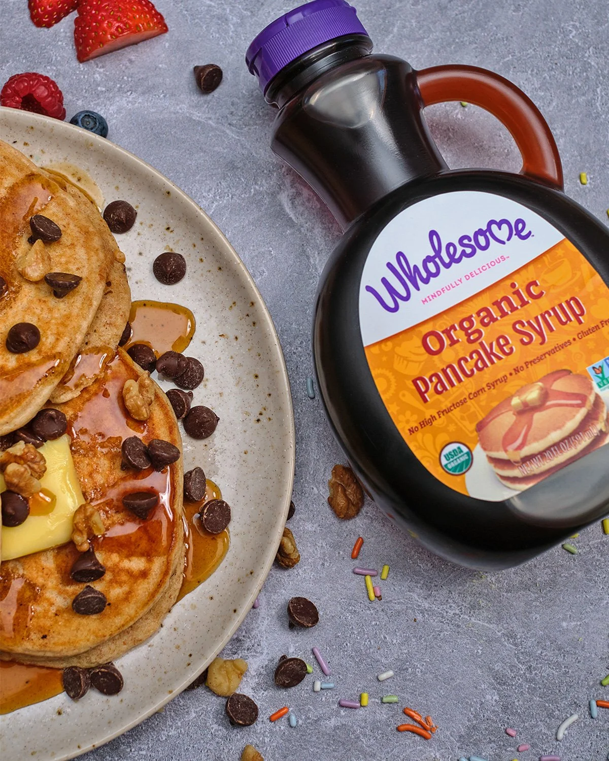 Plate of pancakes topped with chocolate chips, walnuts, butter, and syrup, with scattered sprinkles and crumbs nearby. Bottled organic pancake syrup is nearby.