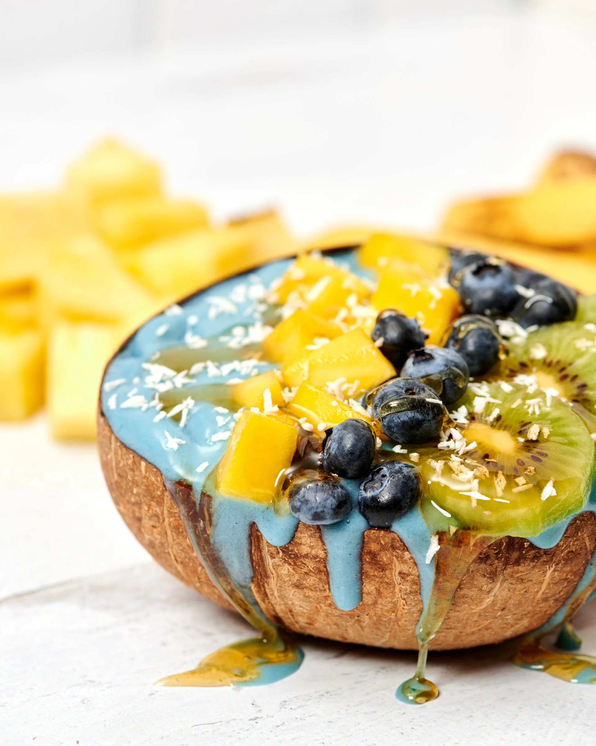 A donut with blue icing, topped with chopped yellow fruit and blueberries, covered in blue and yellow syrup that drips down the sides.