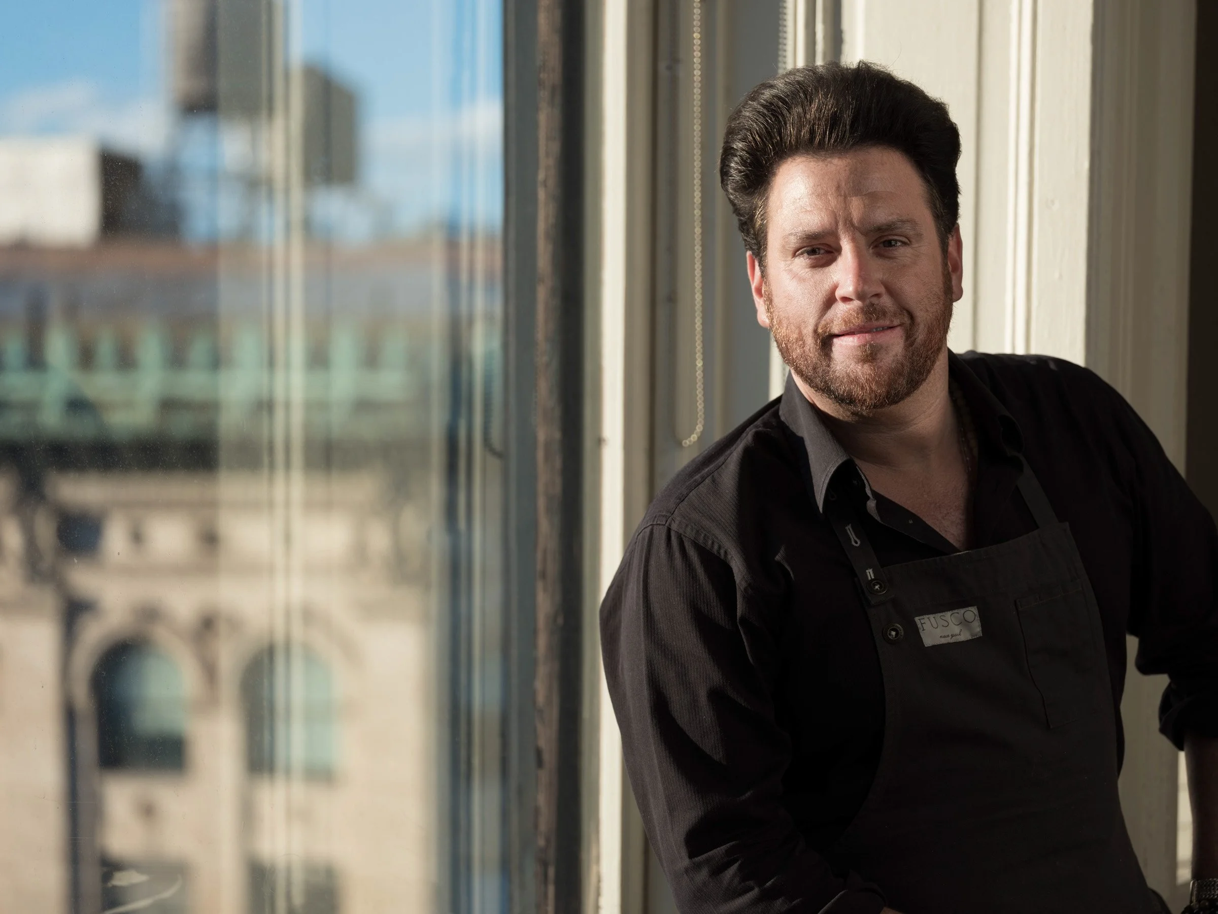 A man with dark hair and a beard, wearing a black shirt with a name tag that says "FUSCO," smiling and leaning against a wall near a large window.
