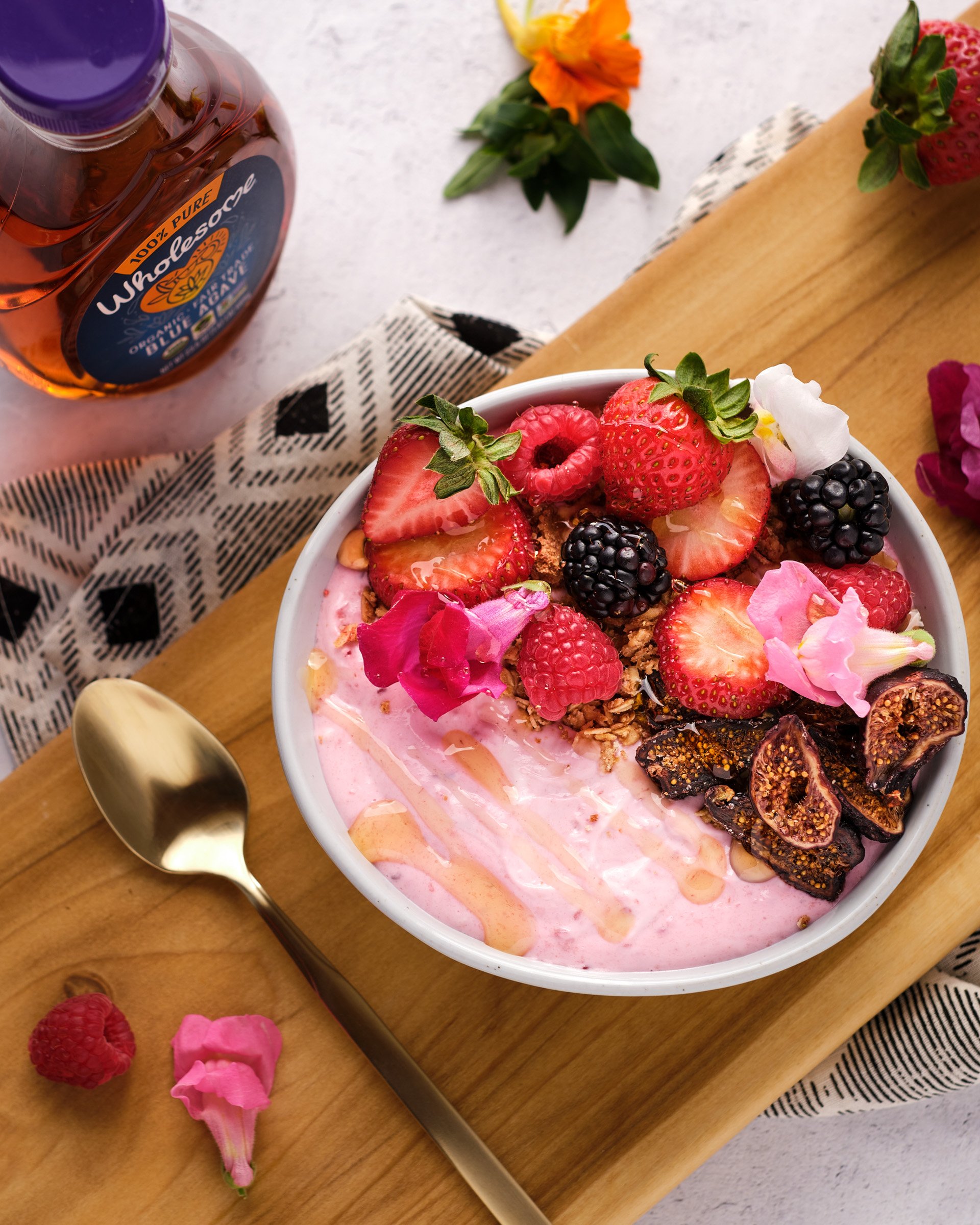 A bowl of pink yogurt topped with sliced strawberries, raspberries, blackberries, chopped figs, granola, and edible flowers, set on a wooden cutting board with scattered berries and flower petals, with a gold spoon nearby.