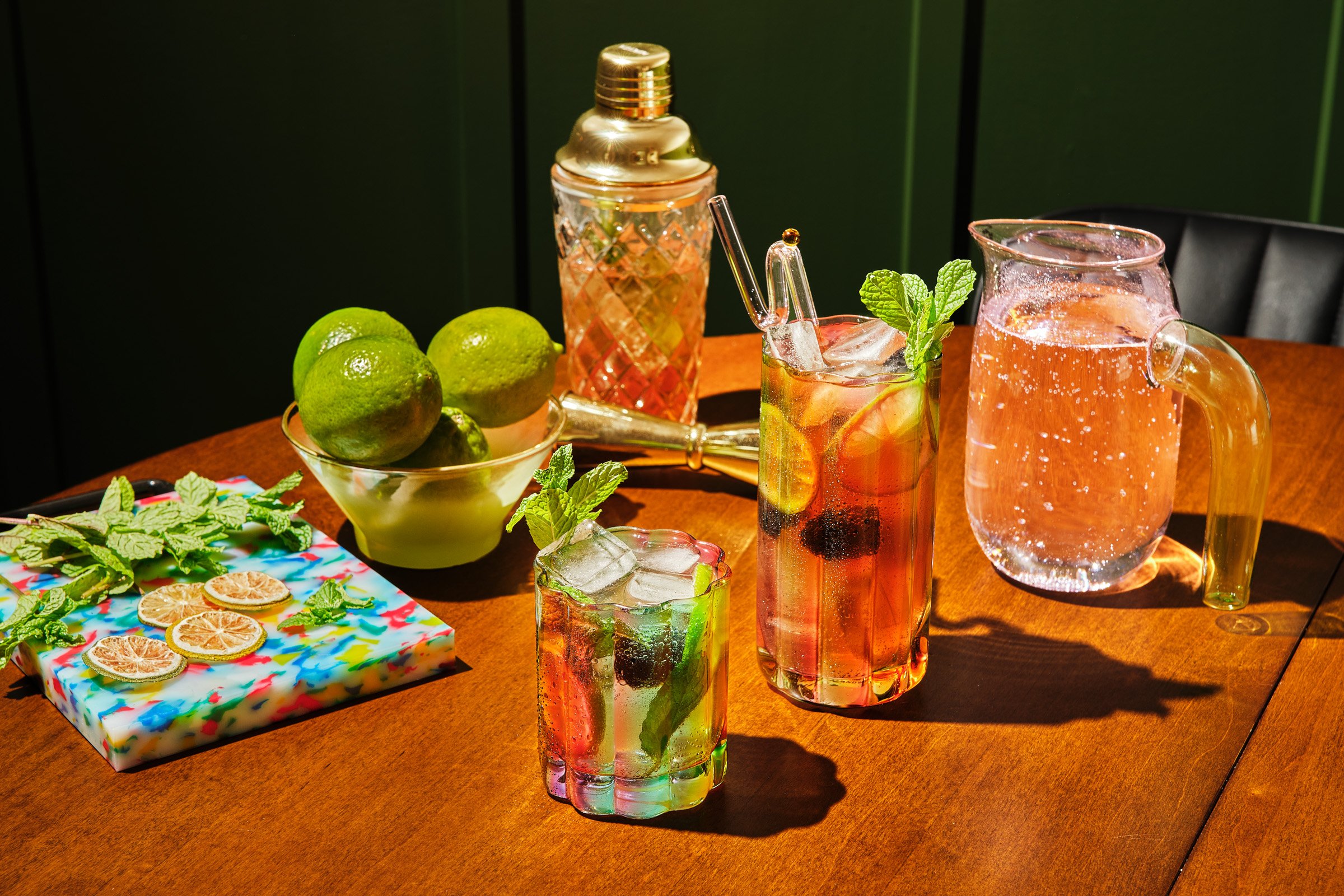 Colorful bar setup with three glasses of mojito cocktails with mint, three limes, a glass jar of pink lemonade, a shaker, and a colorful tray of dried lemon slices and mint leaves on a wooden table against a dark green background.