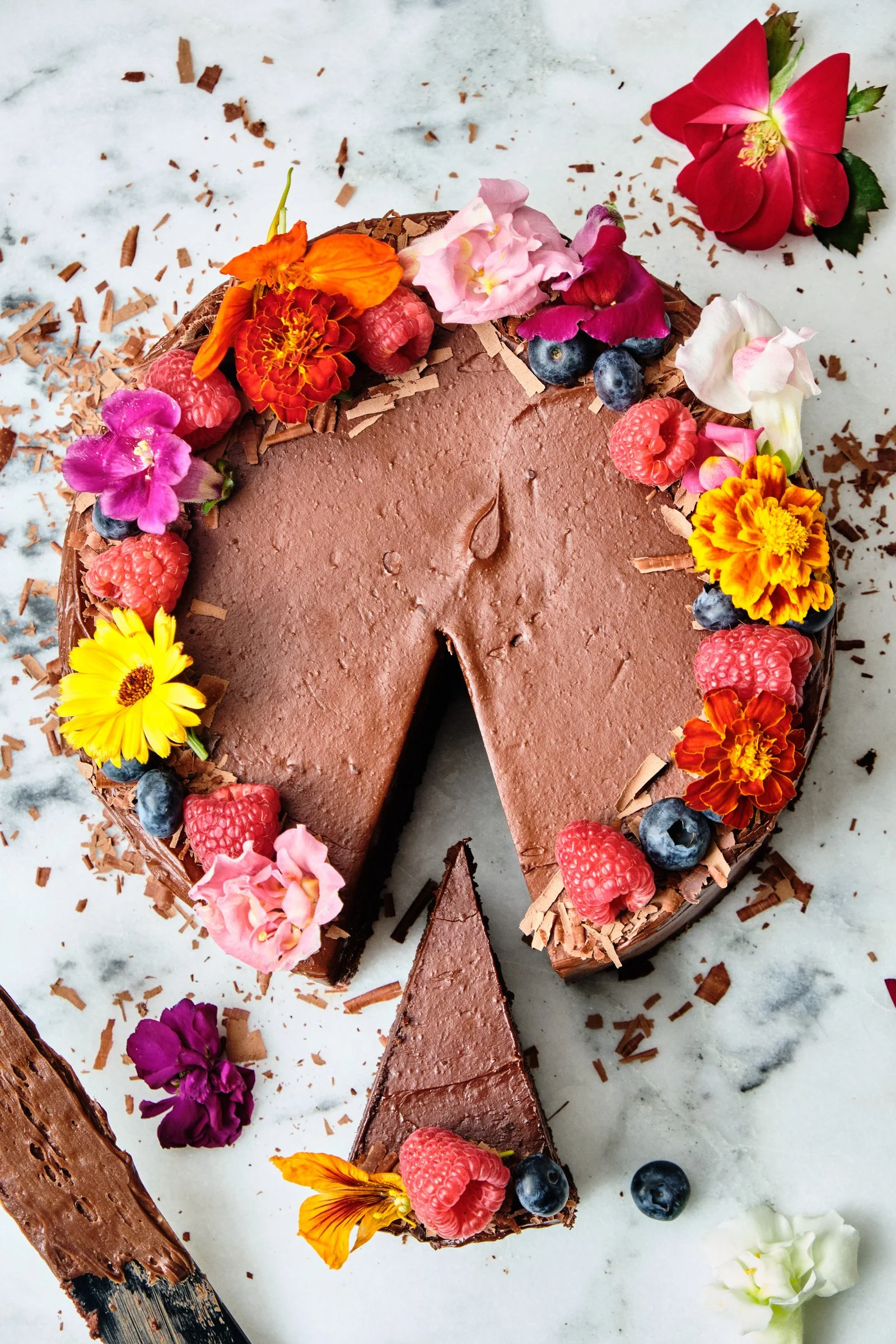 Chocolate cake topped with colorful edible flowers, raspberries, and blueberries, with chocolate shavings around the edges, on a white marble surface with scattered chocolate shavings and flowers.