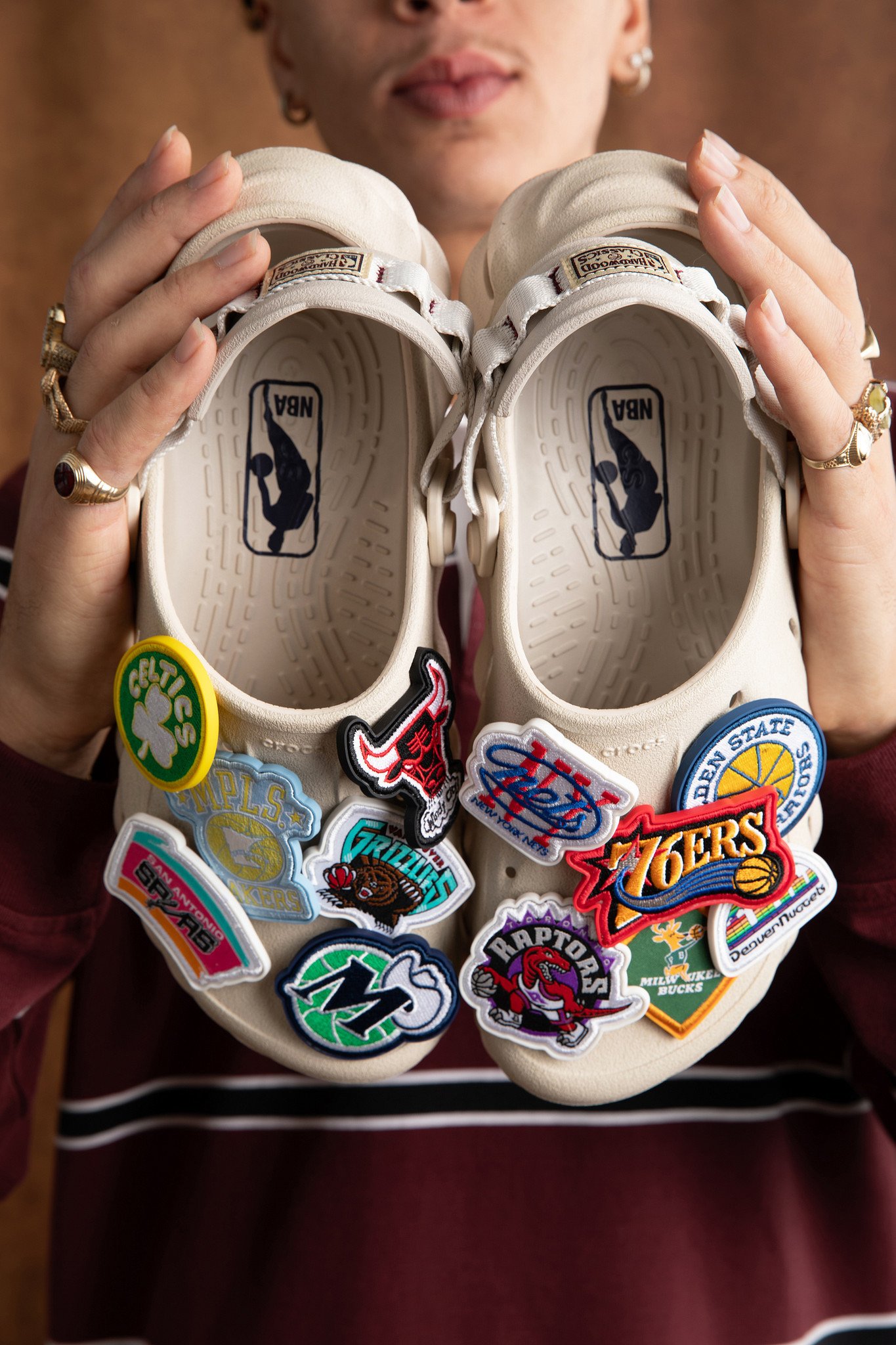 Person holding a pair of white sneakers decorated with various colorful team logos and patches, including NBA teams.