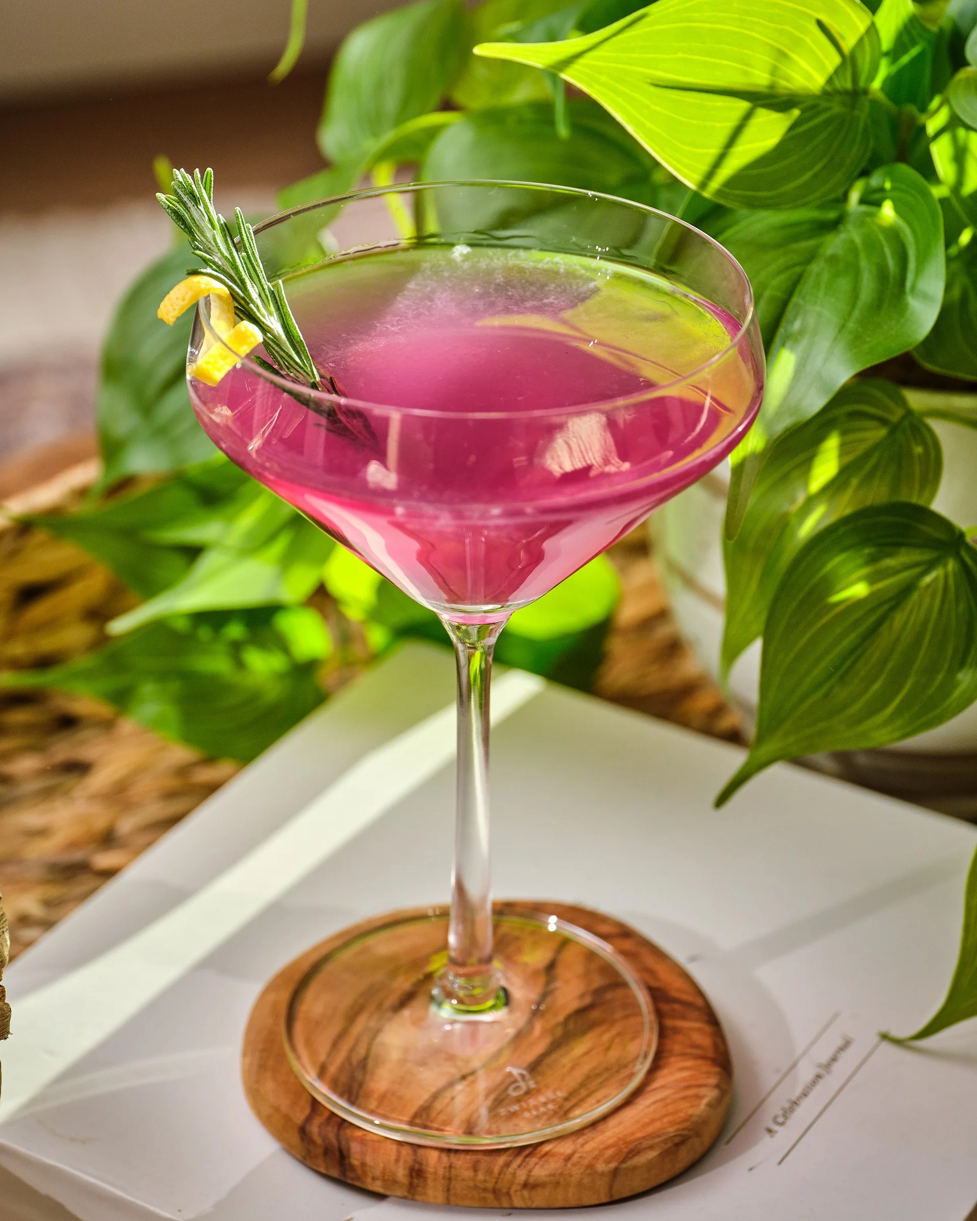 A pink cocktail in a martini glass garnished with a sprig of rosemary and lemon peel, placed on a round wooden coaster with green leaves in the background.
