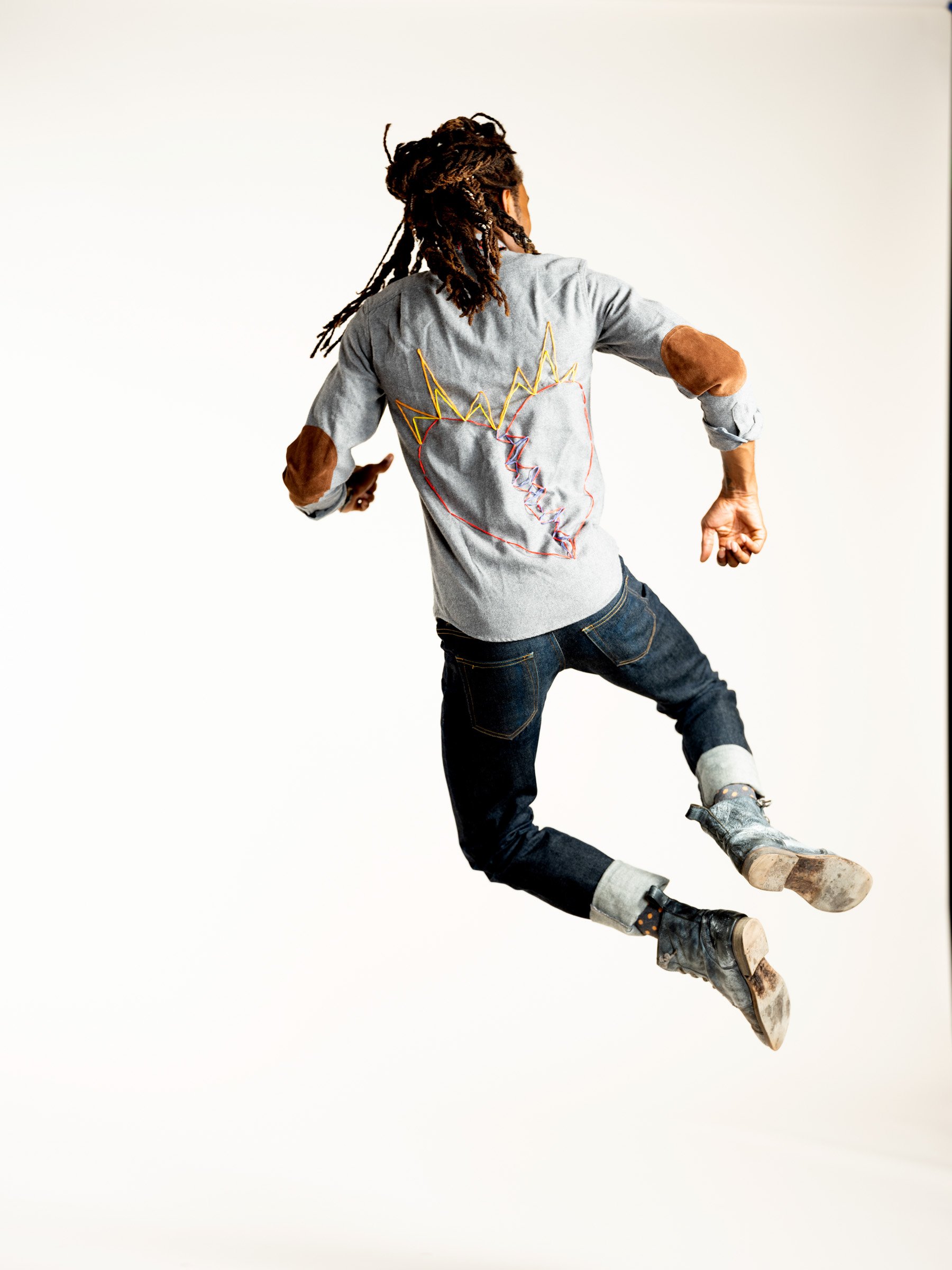 Person with dreadlocks wearing a gray shirt with a heart and crown design, flying in mid-air against a plain white background.