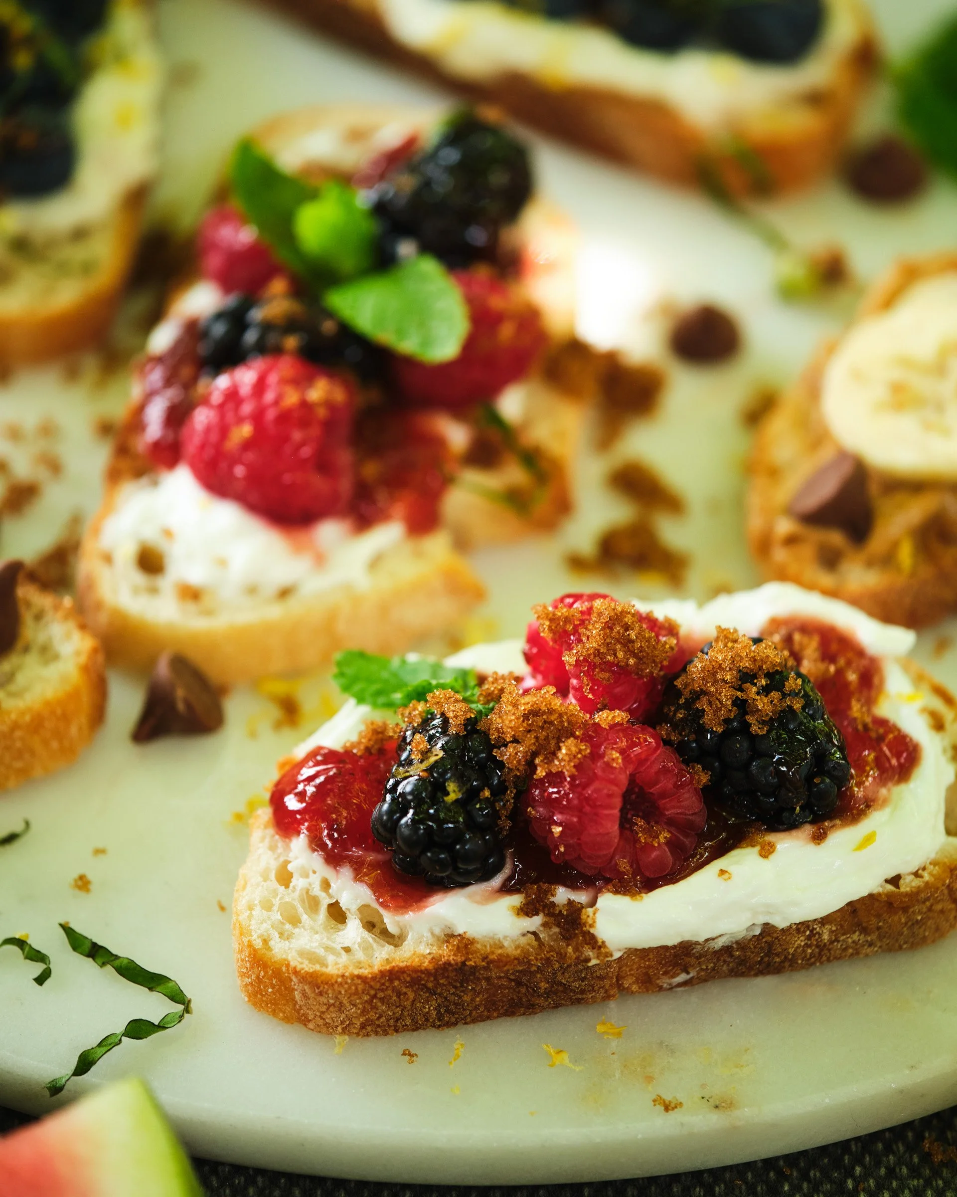 Close-up of a piece of toast with cream cheese, topped with mixed berries, mint leaves, and a sprinkle of crushed cookies or graham crackers.