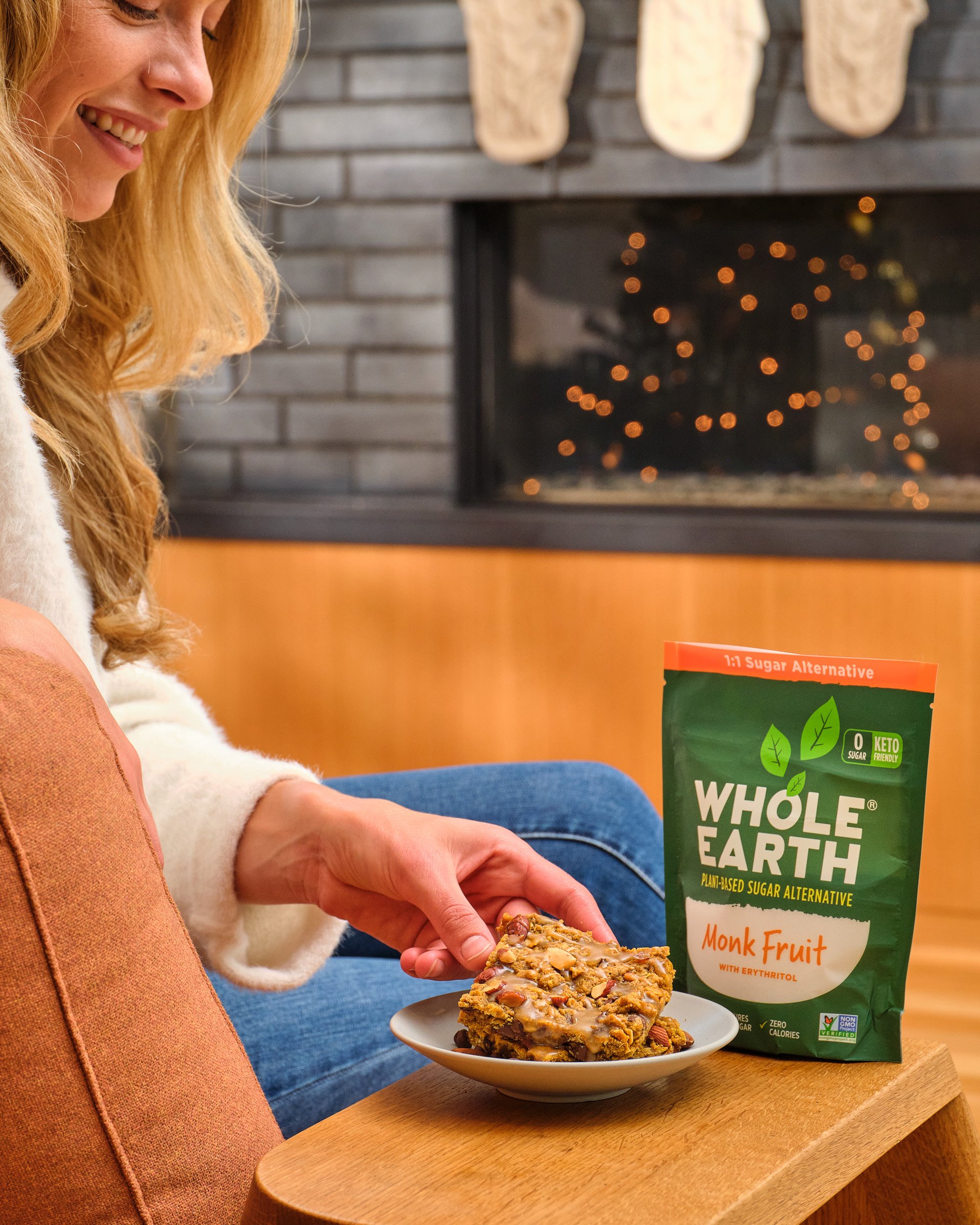 A woman with long, curly blonde hair smiling and holding a granola bar with nuts and chocolate, sitting at a wooden table. In the background, there is a black fireplace with blurred orange bokeh lights and a green Whole Earth Foods package on the tab