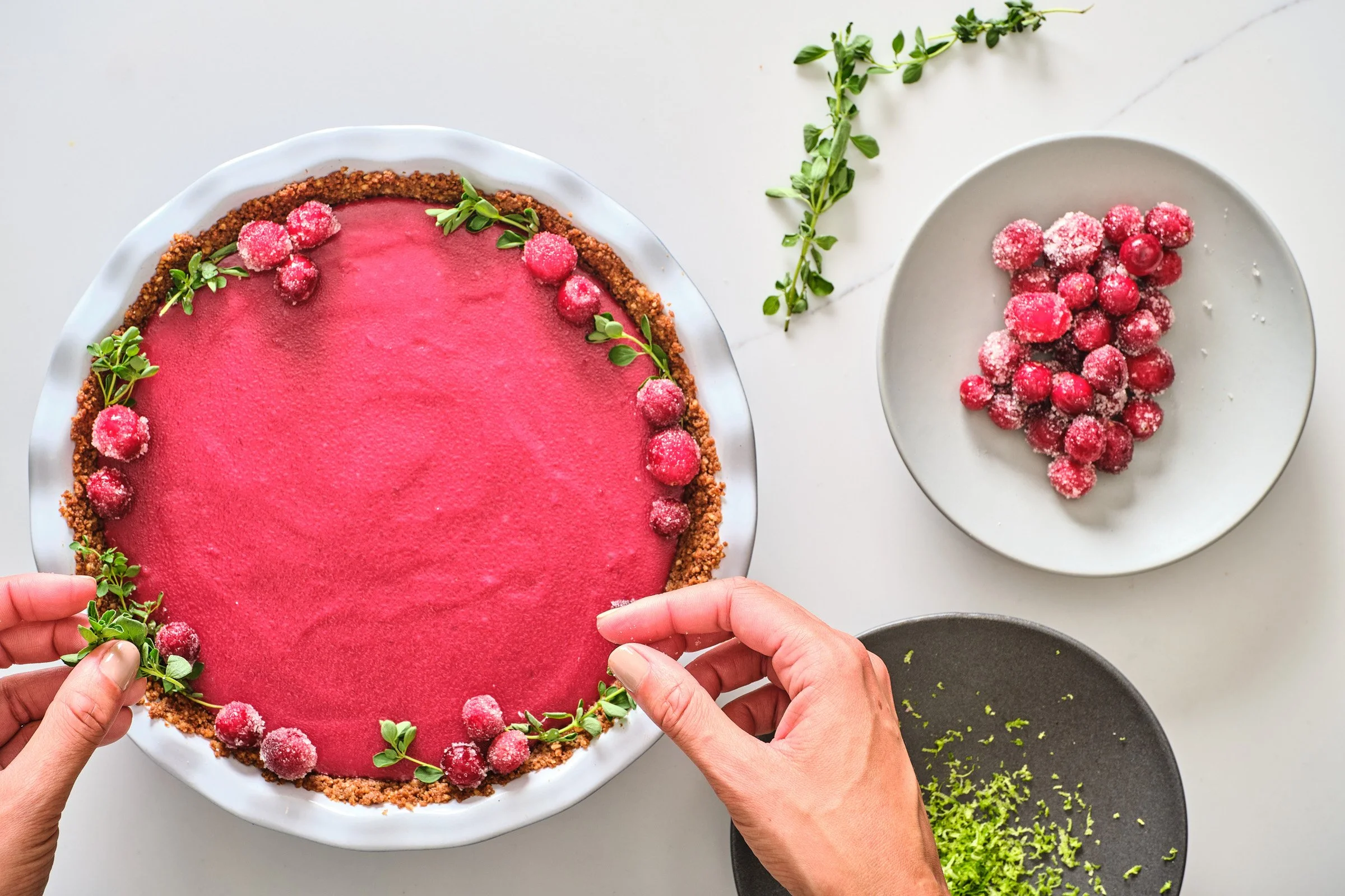 Person decorating a pink berry tart with fresh berries and herbs on a white plate, surrounded by plates of berries and grated lime zest.