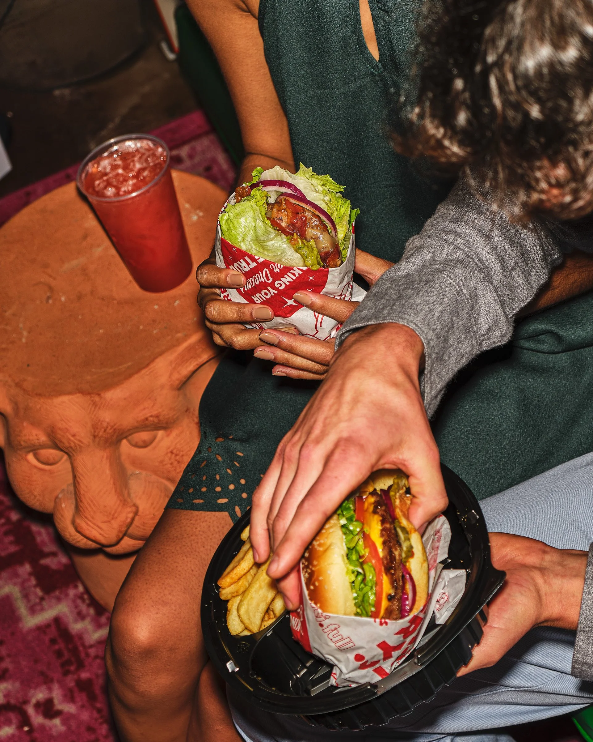 A person sitting on a chair holding a tray with a burger and fries, and another person next to them with a drink in a tall glass.