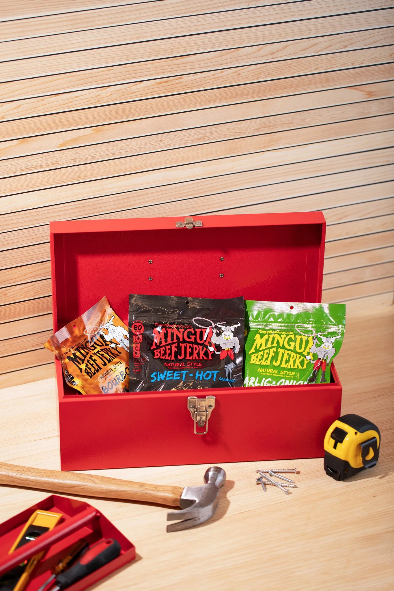 Open red box with packages of beef jerky labeled 'Mingu Beef Jerky' with flavors like 'Sweet Hot' and 'Garlic Onion'; wooden workbench with tools including a hammer, tape measure, screws, and a screwdriver.