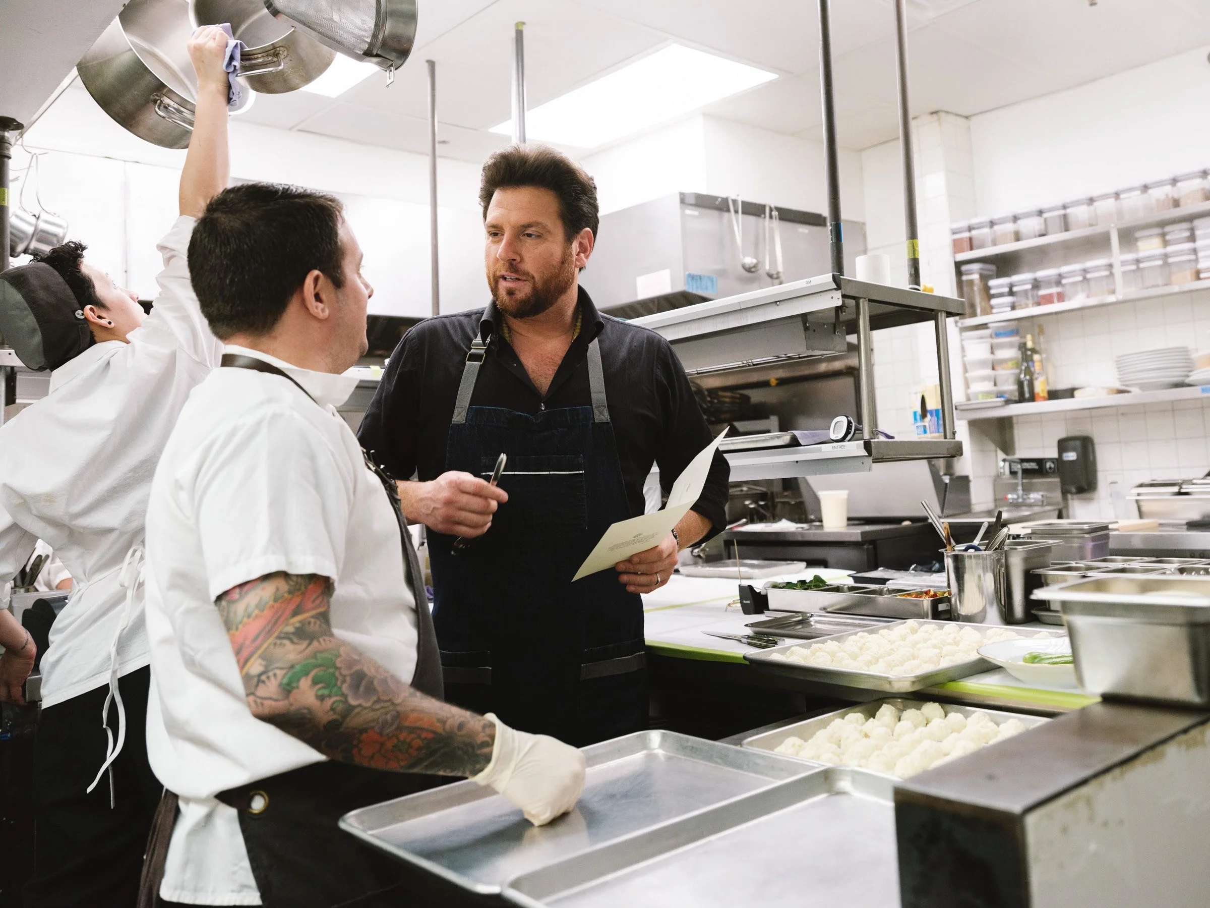 Two chefs in a commercial kitchen talking, one with tattoos on his arm, the other holding a menu or orders, with various kitchen utensils and food ingredients around them.