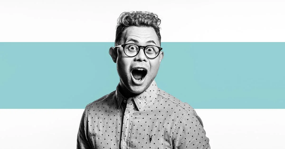 A black-and-white photo of a surprised young man with glasses, wearing a polka-dot shirt, against a background divided into white and blue horizontal sections.