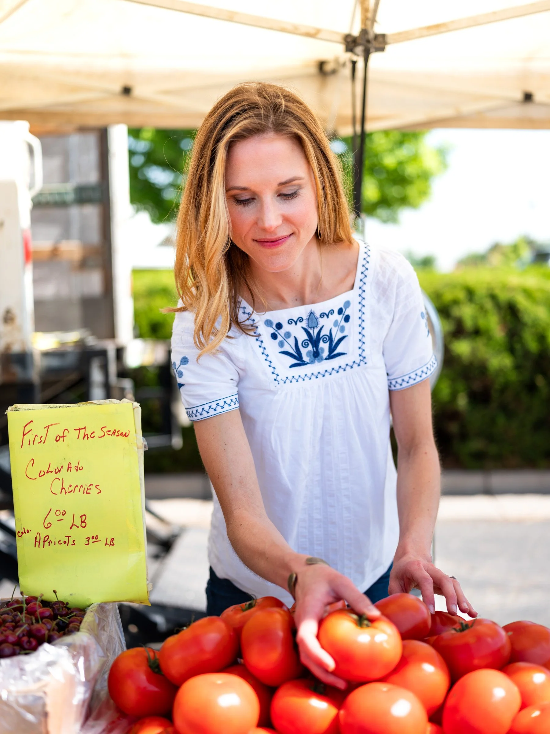 A woman with red hair and a white embroidered shirt is at an outdoor farmers market, selecting ripe tomatoes from a display. There is a yellow sign with red writing listing prices for cherries and apricots. In the background, there are green trees an