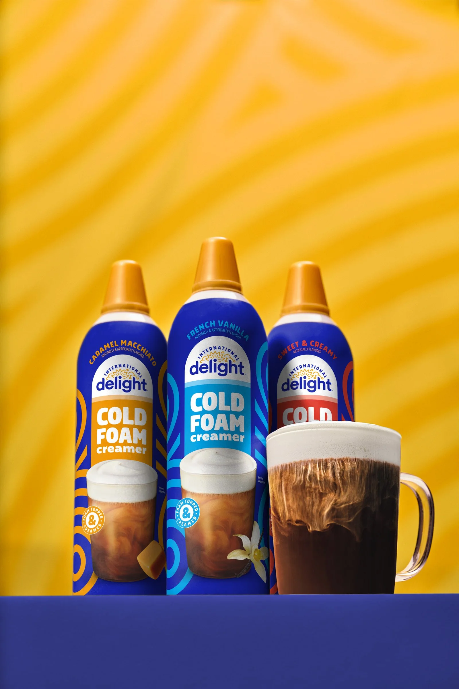 Three cans of delight cold foam creamer in caramel, French vanilla, and sweet and creamy flavors, with a foamed coffee drink and a glass cup of coffee in the background, all against a yellow background.