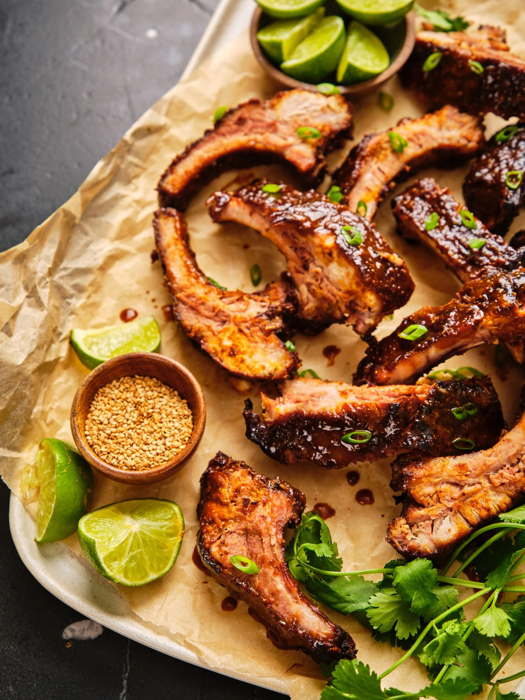 Grilled pork ribs garnished with green onions and cilantro, served with lime wedges, sesame seeds, and a small dish of barbecue sauce on a round white plate lined with parchment paper.